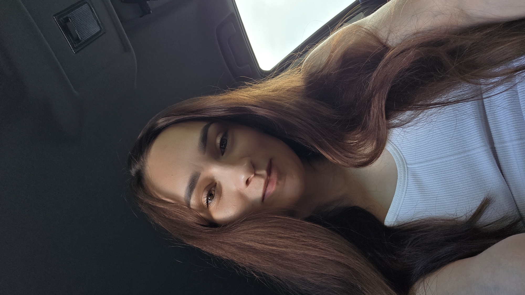 A young woman with long, wavy, brown hair is captured in a close-up, slightly angled shot. She is wearing a white ribbed tank top and appears to be inside a vehicle, as indicated by the dark interior lining and a window visible on the right side of the frame. The lighting suggests she is outdoors, possibly during the daytime, with sunlight catching her hair and casting subtle shadows on her face. Her expression is a gentle smile, with her gaze directed slightly off-camera. The background is dark and out of focus, providing no specific environmental details of Bulgakovo, Russia.