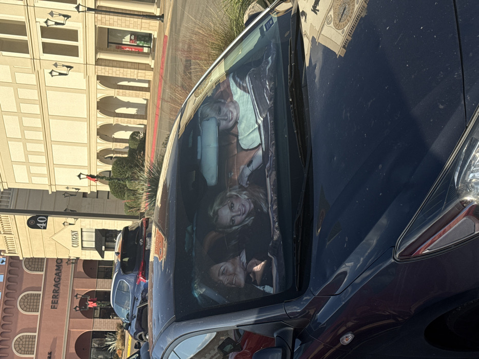 Three smiling women are seen inside a dark car through the windshield. The reflections in the windshield show buildings with retail stores like Ferragamo and Citizen. The car is dark blue and parked on a sunny day. Other cars are parked nearby. The scene suggests a shopping area with multiple storefronts. The light and reflections add depth to the composition.