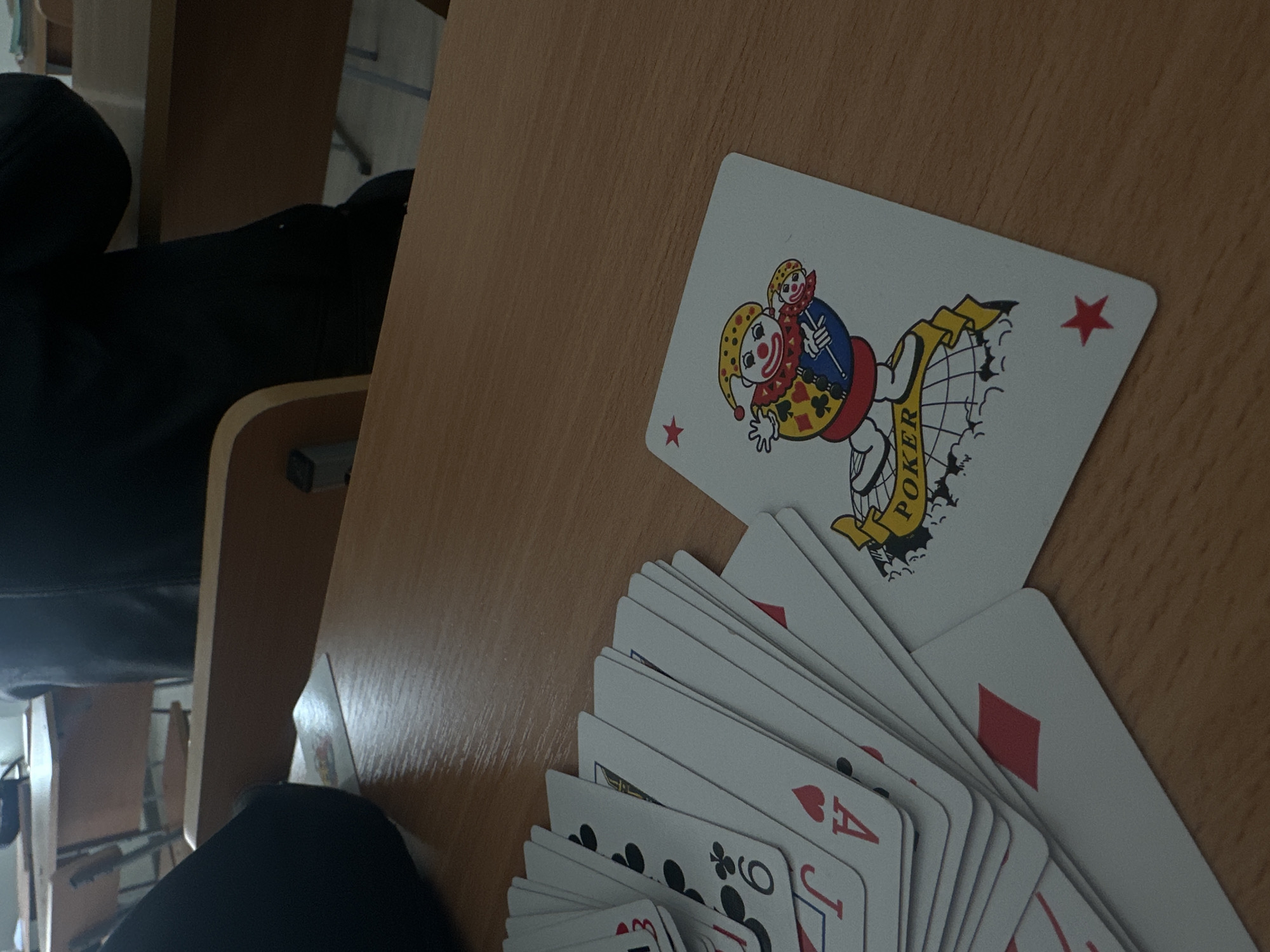 The image captures a close-up, slightly angled view of a wooden desk or table surface. The primary subjects are a deck of playing cards. One Joker card is prominently displayed, lying face up on the table. It features a colorful clown figure holding a smaller clown figure on a stick, with a banner that reads "POKER" vertically. A red star icon is visible in two corners of this Joker card.
Adjacent to the Joker card, a fan of other playing cards is spread out. Partially visible cards include an Ace of Hearts, a Jack of Clubs, a 9 of Clubs, and a card showing a red diamond suit. The cards appear to be standard playing cards.
The activity suggested is someone handling or arranging playing cards, possibly for a game or sorting.
In the background, blurred elements suggest an indoor setting with additional wooden furniture, possibly chairs and desks, reminiscent of a classroom or office environment. A dark, out-of-focus object, possibly an arm or leg covered in dark clothing, is visible in the bottom left, and another dark object appears in the upper left. The lighting is somewhat dim and natural, possibly coming from a window or overhead fixture. No specific time of day or weather can be determined. No emotions are discernible.
