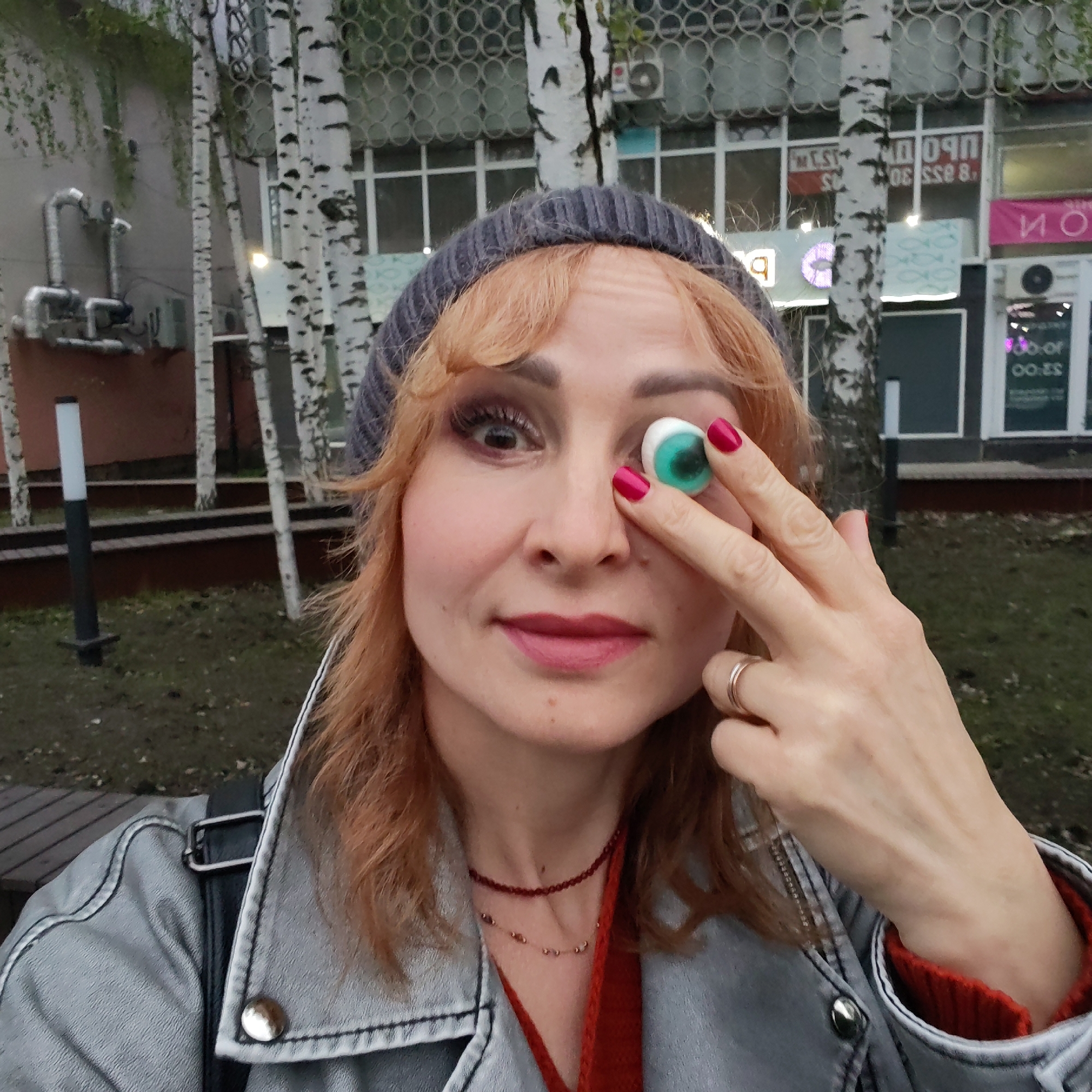 A medium shot depicts a woman holding a candy eyeball in front of her left eye. She has red-blonde wavy hair and is wearing a gray knit hat and a gray jacket over a red sweater. She has red lipstick and matching nail polish on her right hand. A gold ring adorns her ring finger. Around her neck, she wears a maroon beaded necklace and a gold chain.
The background features birch trees, a building with barred windows, and signs written in Russian, indicating the location is likely in Russia. The lighting suggests it is daytime, though the weather appears overcast. Further information suggests this image was taken in the city of Chaikovsky, Russia.