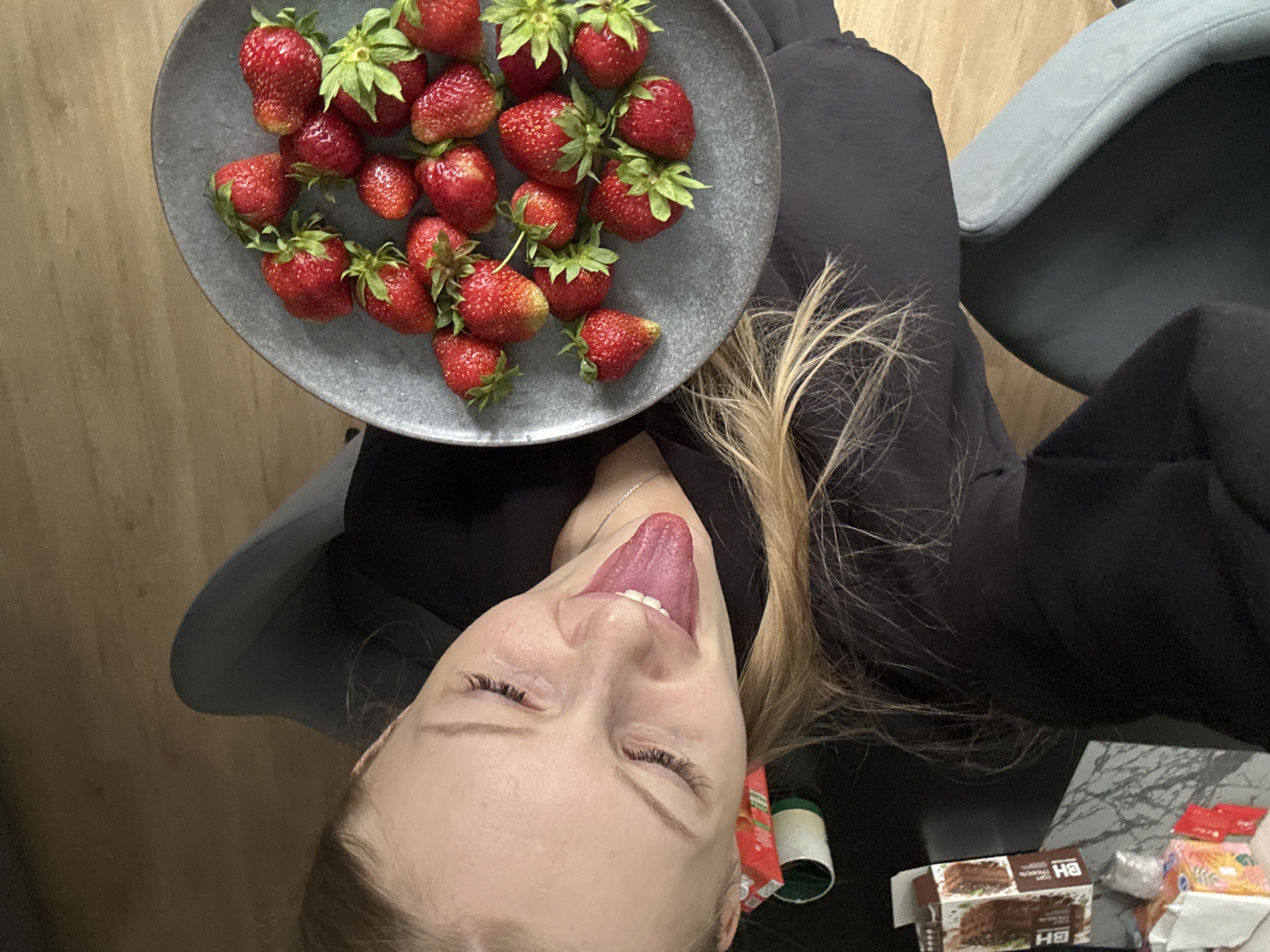 A woman is captured from a high angle, looking up at the camera with her eyes closed and her tongue sticking out, positioned at an unusual angle to her face. She is wearing a black top and seated in a chair. To her right, a gray plate holds a pile of fresh strawberries. The background consists of a wooden floor. To her left, on a table, are several items including a small box, a roll of tape, and some other packaging. The scene appears to be indoors, possibly in a residential setting, in the city of Perm, Russia.