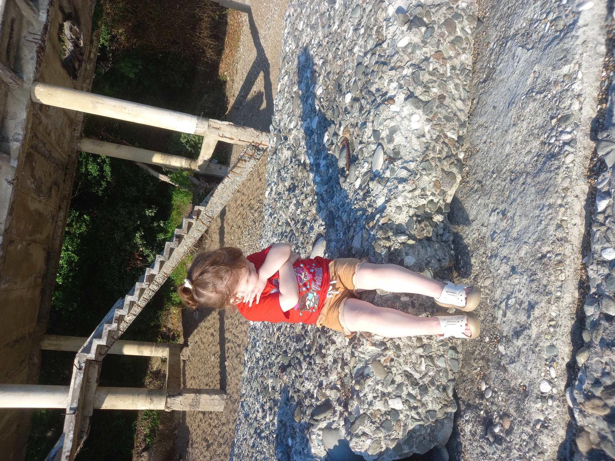 A young girl with brown hair tied in a bun is sitting on a large concrete structure composed of many rocks. She is wearing a red t-shirt with graphics and tan shorts, and white sandals. Her arms are crossed and she is looking down. The concrete structure is on a beach, with sand and smaller pebbles around it. In the background, there is a partially ruined concrete structure with columns and stairs, surrounded by green foliage. The overall setting appears to be an outdoor, somewhat neglected, coastal area. This location is identified as Gwandra, Abkhazia.