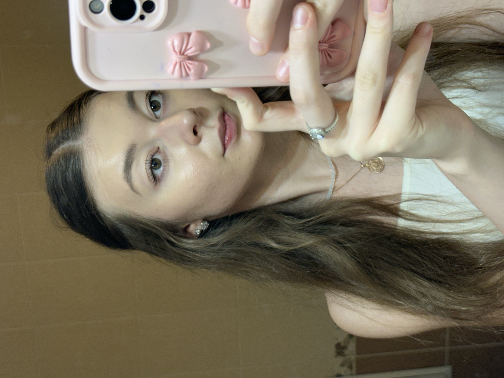 The image is a selfie taken by a young woman with long, wavy brown hair. She is wearing a white top, a delicate necklace with a pendant, a ring on her right hand, and diamond stud earrings. Her right hand is holding a pink phone case adorned with multiple pink bows. The background appears to be a tiled bathroom wall. The image does not provide any information about the location in the city of Kazan, Russia.