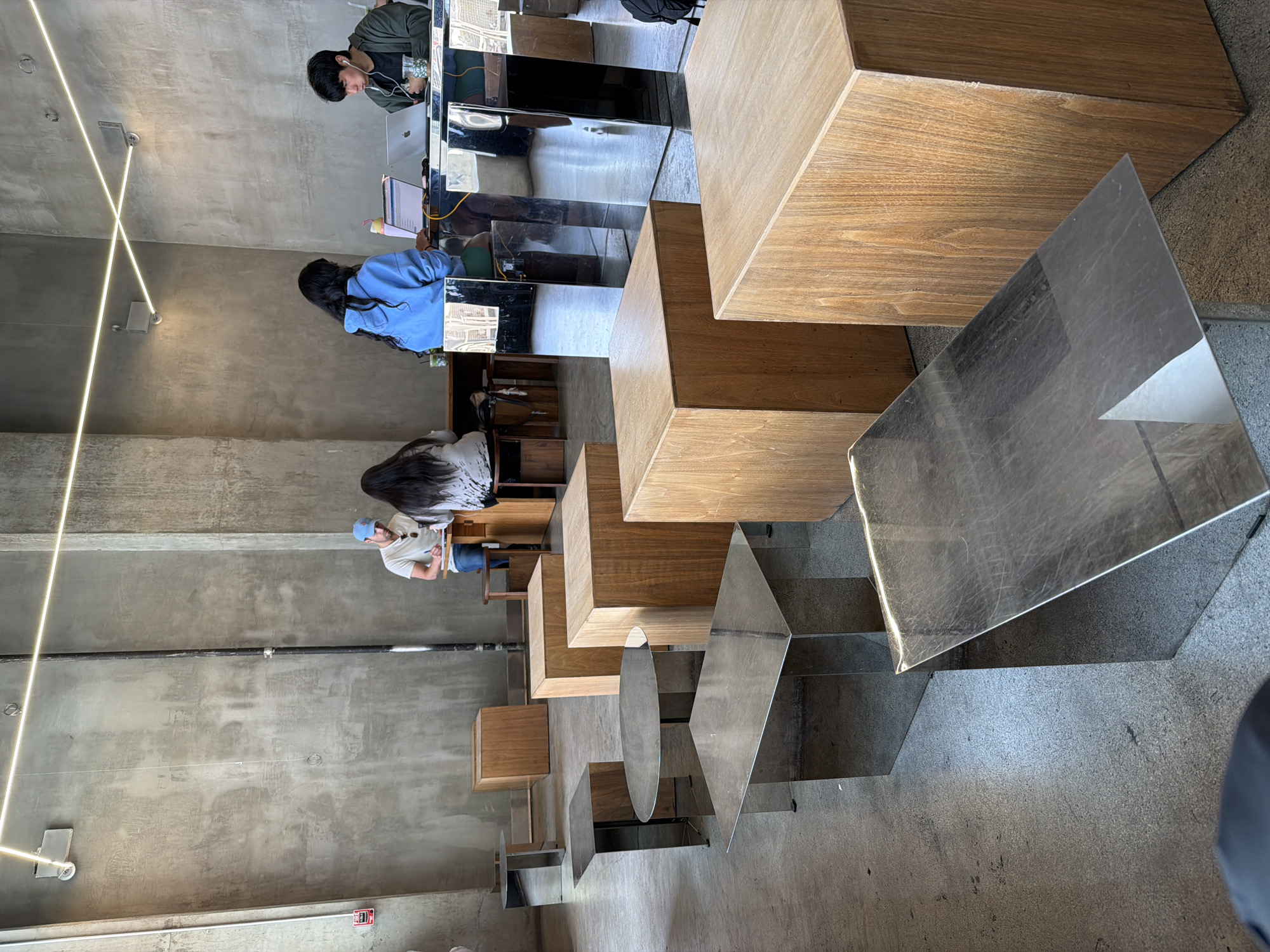 The image shows an interior space, seemingly a cafe or coworking area, in an urban setting. The photo has been taken from a slightly elevated vantage point, looking down towards the seating and working areas.
The scene features multiple people engaged in various activities, presumably working or studying. There is a man in a green shirt using a laptop at a desk. Another individual is wearing a blue top. Another person with long dark hair is also seated at a workspace. Lower down in the image, another individual in a white shirt is seated at a desk.
The furniture consists of modular, geometric wooden and mirrored tables.
The environment is characterized by concrete walls. Lighting fixtures are visible on the ceiling.
The location appears to be within the city of Los Angeles, United States of America.