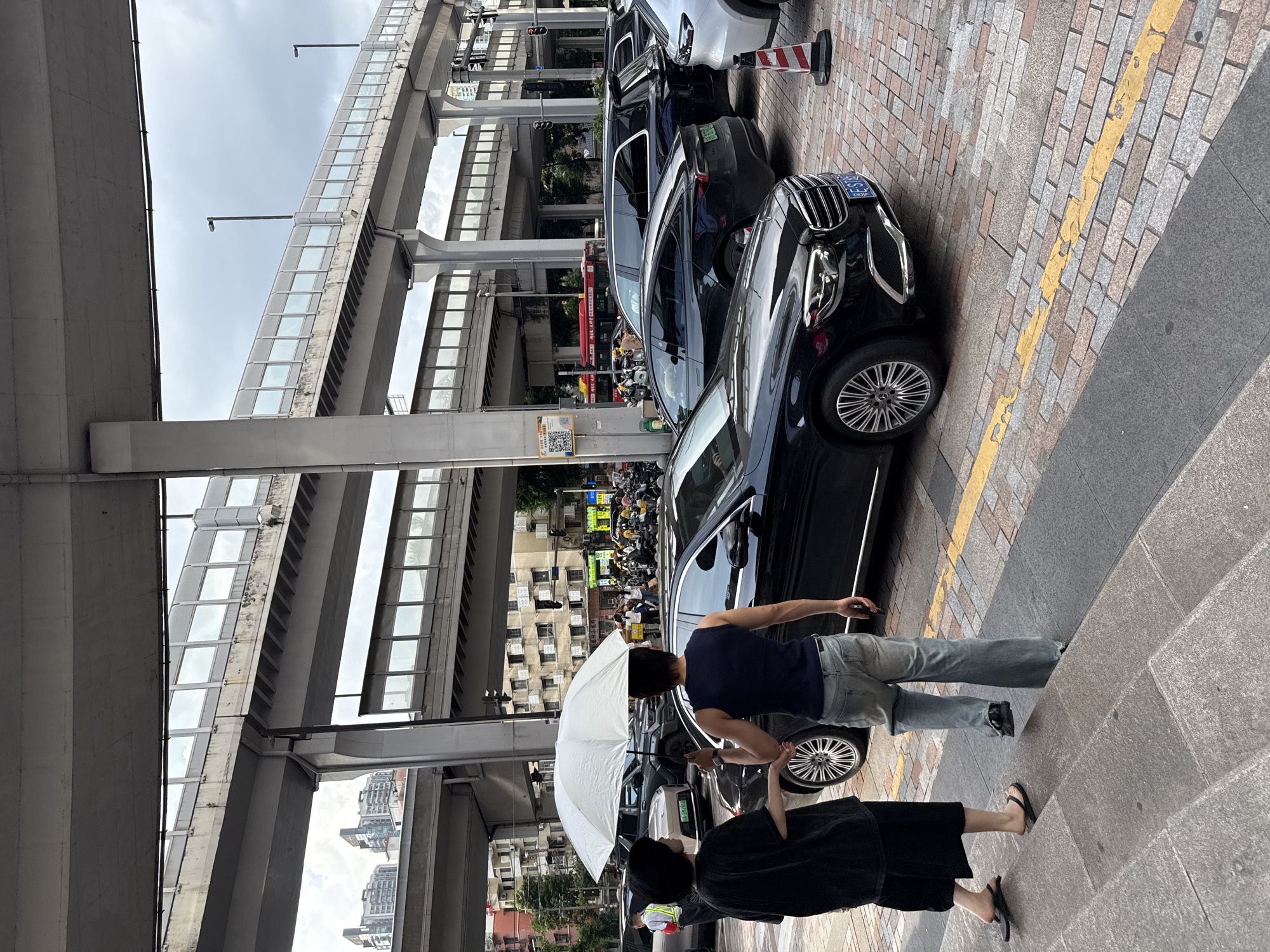In Haizhu District, China, two individuals are walking on stairs with a white umbrella overhead. The woman, on the left, is wearing a black outfit and black flip-flops. The man, on the right, is wearing a dark blue sleeveless shirt and loose-fitting blue jeans. They are walking towards a black Mercedes-Benz sedan parked on a cobblestone street. Several other vehicles, including SUVs and motorcycles, are also visible. Elevated highways, supported by concrete pillars, dominate the background, with buildings and traffic lights also present. The sky is overcast.