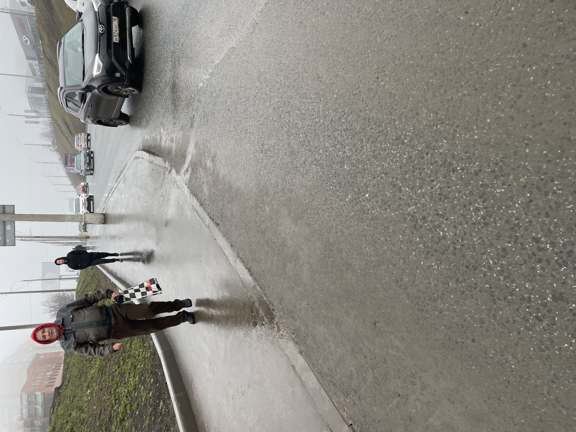 The image captures a street scene in Izhevsk, Russia, on what appears to be a wet or rainy day. The location is along a road, with several cars visible in the background, including a dark SUV in the upper-left corner with a license plate that begins with "M 924 OC 18." There are buildings visible in the background, shrouded in fog or haze. A sign that reads "TENET" is visible in the background.
In the foreground, a man wearing a red head covering, a brown jacket, and brown pants stands on the sidewalk. He is holding a black and white checkered bag or umbrella. Behind him and further up the sidewalk, another person in dark clothing is walking in the same direction. The sidewalk is wet, reflecting the surroundings.
