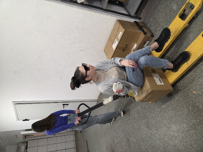 The image depicts an indoor scene in the city of Varna, Bulgaria, featuring two females and a manual pallet jack. In the foreground, a young female is seated on a large brown cardboard box, which rests on the yellow forks of the pallet jack. She wears a grey cardigan over a beige top, light blue distressed jeans, and black sneakers. Her dark hair is secured in a messy bun with a black scrunchie, and she wears large dark sunglasses. She holds a white mug in her right hand and a small purple object in her left; a watch or bracelet is visible on her left wrist. The cardboard box serving as her seat displays a white label with a barcode and 'EAC' text. Behind her, a second young female, viewed from the back, has long dark hair and wears a blue long-sleeved top, light blue jeans, and light-colored sneakers. She holds the black handle of the pallet jack with both hands, appearing to operate it. To the right, two additional brown cardboard boxes are stacked against a plain white wall; one of these boxes also displays an 'EAC' label. A partial metal shelving unit is visible to the far right. The left background features two white doors with black hardware and grey metal lockers. The floor is grey and textured throughout the space.
