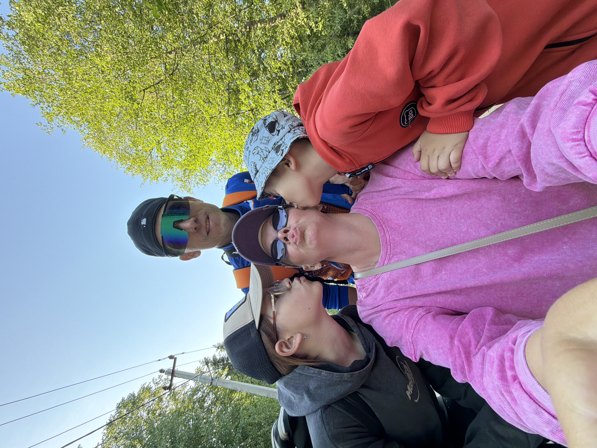The image shows a selfie of a family of four with green trees and a clear blue sky in the background. It seems to be taken from a low angle, tilting upward.
From left to right:
A man wearing a black baseball cap and iridescent sunglasses stands to the left. He wears a blue and orange athletic jacket.
A young girl with a baseball cap is below and kissing the woman.
A woman in a pink t-shirt wearing sunglasses is in the middle and being kissed by two children.
A young boy with a dinosaur patterned bucket hat and wearing a red sweatshirt is kissing the woman’s cheek.
Telephone poles and power lines are visible behind them, and green foliage from trees fills the upper portion of the background. Given the request, the location is Chaikovsky, Russia.