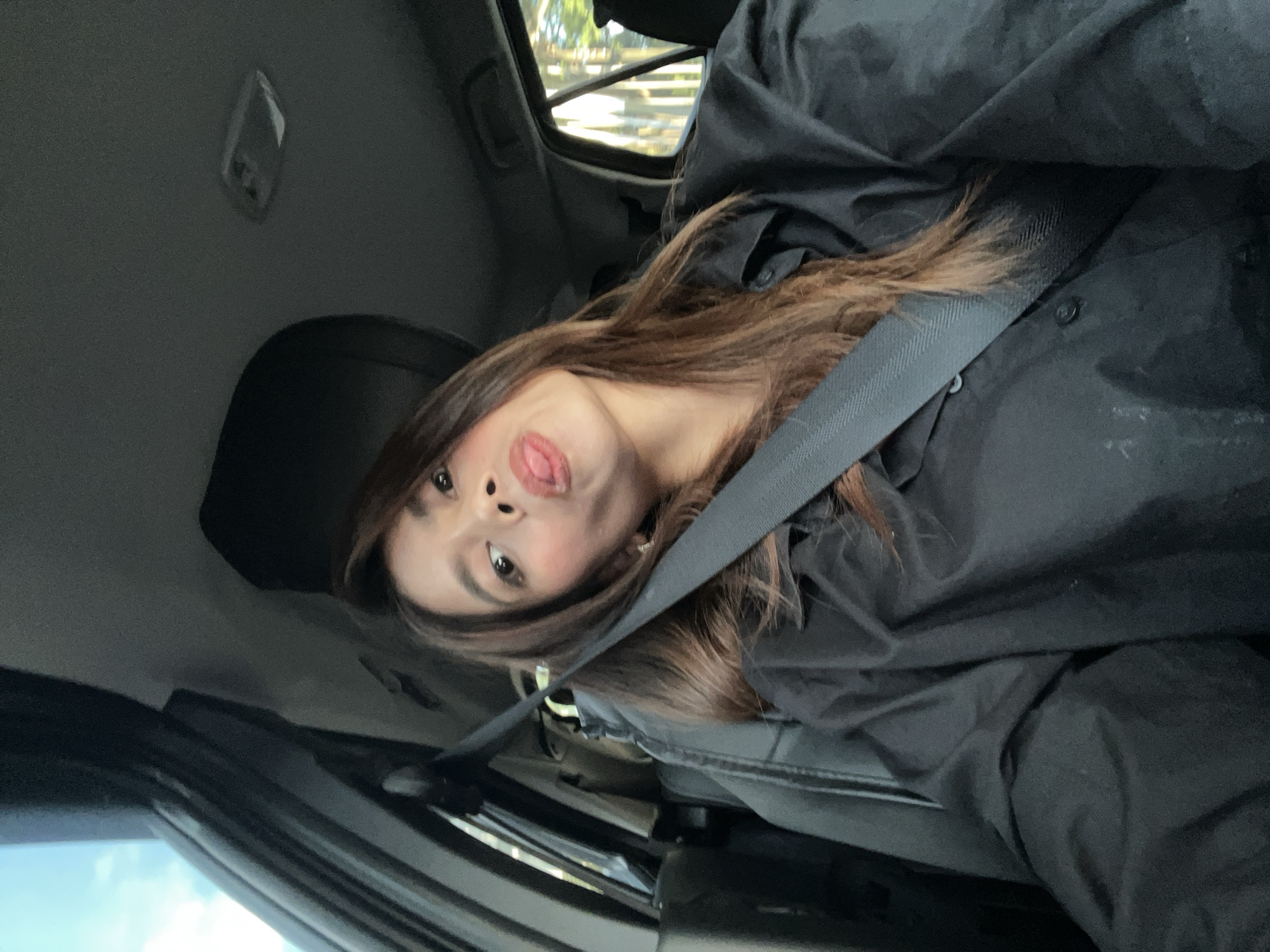 A young adult female is depicted in a selfie taken inside a passenger vehicle, described as being in Davao City, Philippines. She has long, dark brown hair and is wearing a black collared button-up shirt with a dark seatbelt securely fastened across her chest and shoulder. Her face is angled towards the camera, with a playful expression featuring her tongue slightly extended. Her right eye is slightly narrowed, while her left eye is wider open. The car's interior features dark-colored upholstery and headliner, with a white dome light fixture visible on the ceiling. Through the vehicle's windows, blurred daylight and partial views of blue sky, green foliage, and what appears to be a light-colored fence or building are visible, suggesting an outdoor setting during daytime.