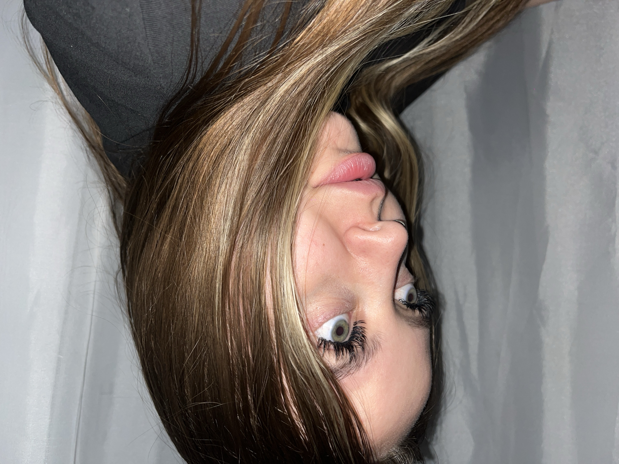 A close-up portrait features a young person, likely female, positioned with their head tilted significantly backward or lying down and looking upwards. Their long, straight to wavy hair, a blend of brown with blonde highlights, frames their face.
The person's eyes are wide open, displaying a direct and intense gaze with light green or hazel irises. Prominent, dark eyelashes, which appear to be false or extended, draw attention to the eyes. Their lips are full, pink, and slightly parted. Part of their nose is visible from an upward angle.
A dark, possibly black, garment is visible around the upper chest and shoulder area. The background is plain and light-colored, appearing as a soft grey or white, possibly a curtain or wall, suggesting an indoor setting.
The image captures a posed moment, focusing on the facial features and the unusual upward angle. There are no specific activities depicted beyond posing for the photograph. No text is visible in the media. While the location context is given as Krasnodar, Russia, there are no visual cues within the image itself to confirm this.