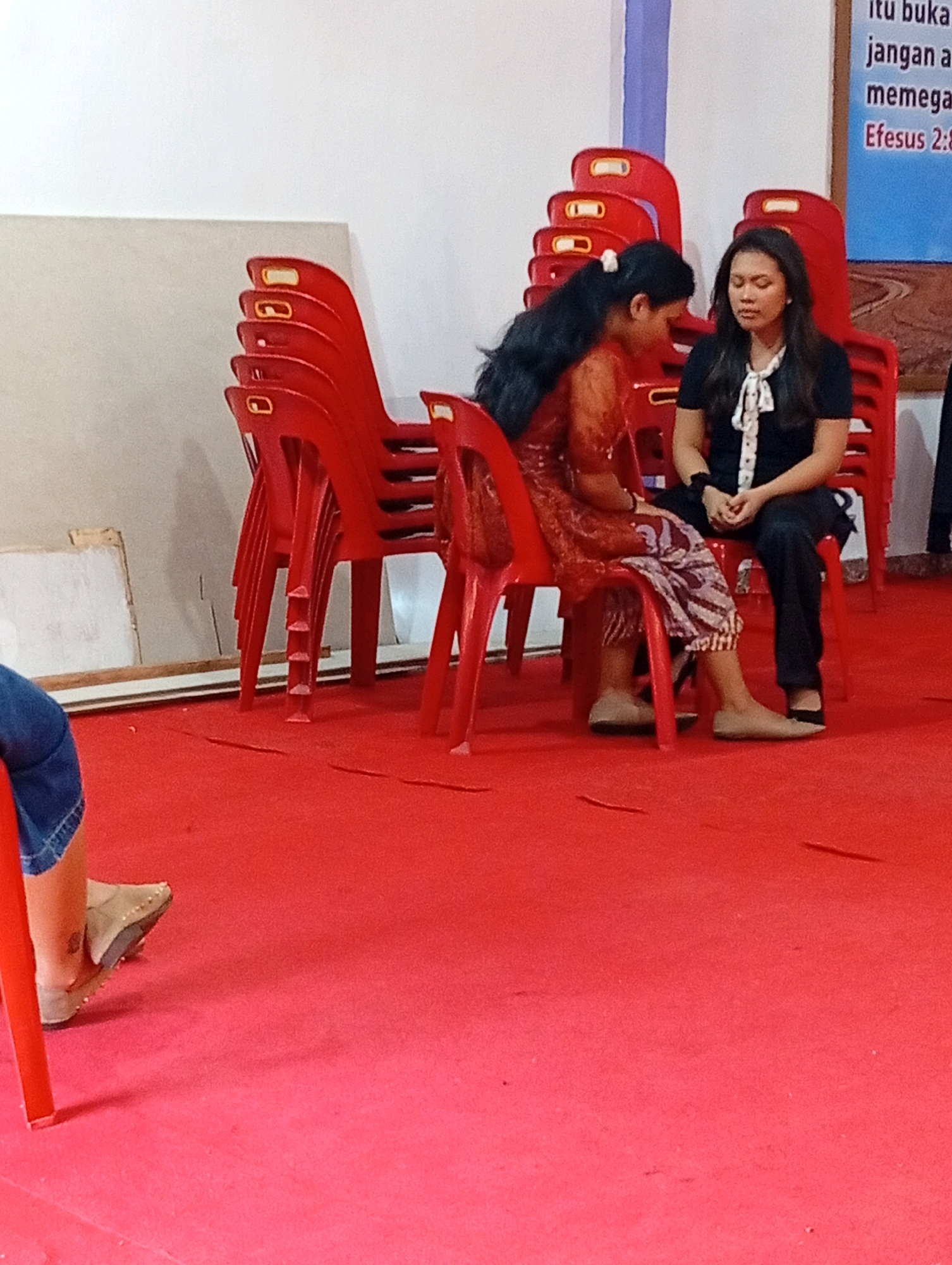 The image depicts two women seated on red plastic chairs in a room with a red carpet. The woman on the left, with long black hair, is wearing a reddish-orange patterned dress and is looking down. The woman on the right, with dark hair, is wearing a black top with a white ascot and black pants, and appears to be looking down with her eyes closed. Behind them, several stacks of red plastic chairs are visible. To the right of the women, a portion of a banner with text in Indonesian is visible. The text mentions "itubuka jangan a memega Efesus 2:8". This text suggests a religious context, likely within a church or similar institution. The presence of stacks of chairs indicates a gathering or event is taking place or has recently concluded. The environment appears to be indoors with plain white walls. The overall scene is static and focused on the two seated individuals. Based on the Indonesian text on the banner, the location is likely in Indonesia. While the specific city is not explicitly stated in the image, the banner text mentioning "Efesus 2:8" (Ephesians 2:8) is a biblical reference commonly used in Christian contexts. Without further context, it's impossible to pinpoint the exact location within the city of Gunungsitoli, Indonesia.