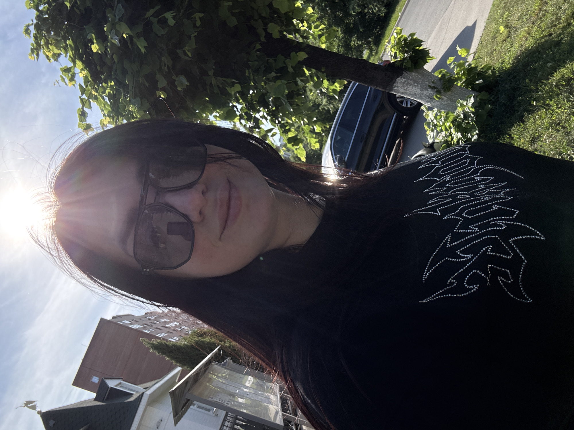 A woman is taking a selfie outdoors. She is wearing sunglasses and a black top with a design made of small, shiny studs. Sunlight is shining from the upper left of the frame. In the background, there is a tree with a whitewashed trunk, a parked car, a street, grass, and buildings. The location appears to be a residential area in a city.