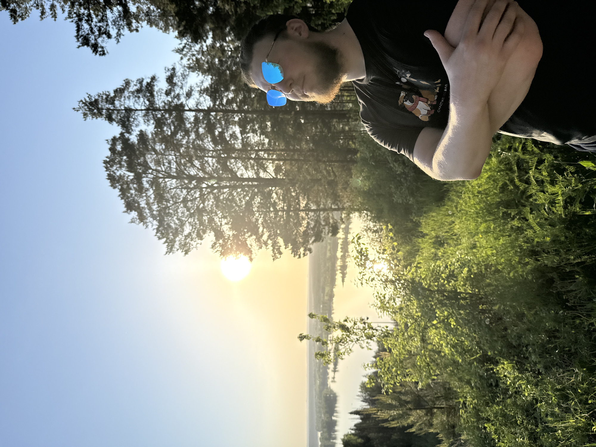 A bearded man wearing blue mirrored sunglasses and a dark-colored T-shirt with a cartoon graphic stands with his arms crossed in an outdoor setting. The man appears to be facing towards the right, with his gaze following the sunset in the background. The scene is bathed in a warm, golden light from the setting sun, which is visible through the trees. There is a body of water, possibly a river or lake, with what seems to be a city or town located on the opposite shore. The surrounding area includes trees and some green vegetation, suggesting a park or natural environment. Given the prompt specifying Noviy, Russia, it can be inferred that the location is somewhere within or near that city, with a vista overlooking a waterway and urban area.