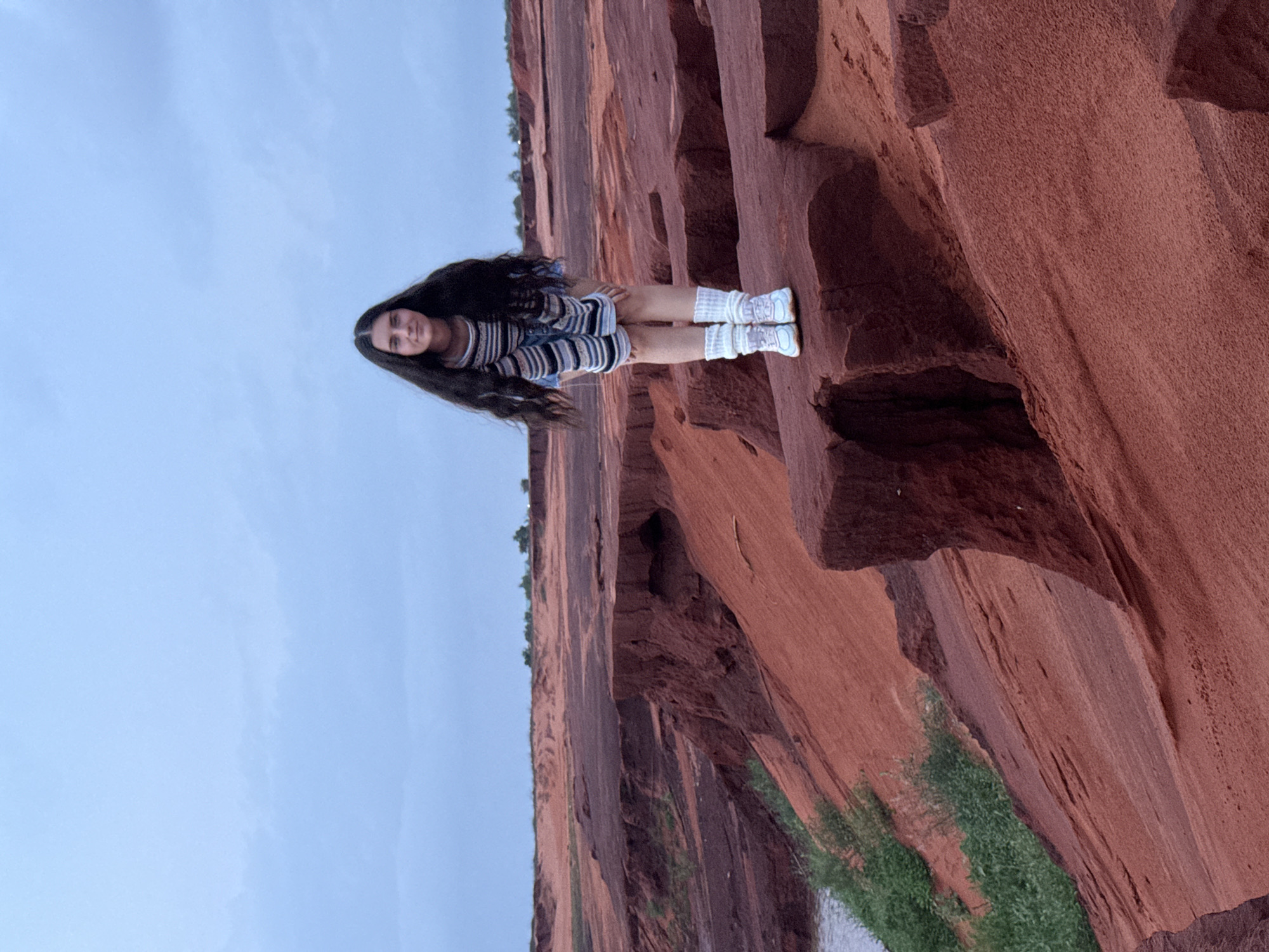A young woman stands prominently on a reddish-brown sandstone ledge, set against a backdrop of similar rock formations and a clear, light blue sky. She possesses long, dark hair that flows over her shoulders and wears a striped long-sleeved top with horizontal black, white, and grey bands, paired with dark shorts. Her attire also includes white athletic shoes and white, ribbed leg warmers. She faces forward, looking towards the viewer with a gentle, serene expression.
The surrounding landscape is dominated by the striking red and brown hues of layered rock, sculpted into cliffs and undulating surfaces. Patches of sparse green vegetation are visible growing on the rock faces and at the base of the formations, adding a touch of contrast to the arid tones. The sky is bright and cloudless, suggesting daytime, possibly late afternoon, given the soft, diffused light illuminating the scene. The distinctive geological features strongly indicate a natural outdoor setting, characteristic of the Red Canyon or Fairy Stream areas in Mui Ne, Vietnam. No text is visible in the image.