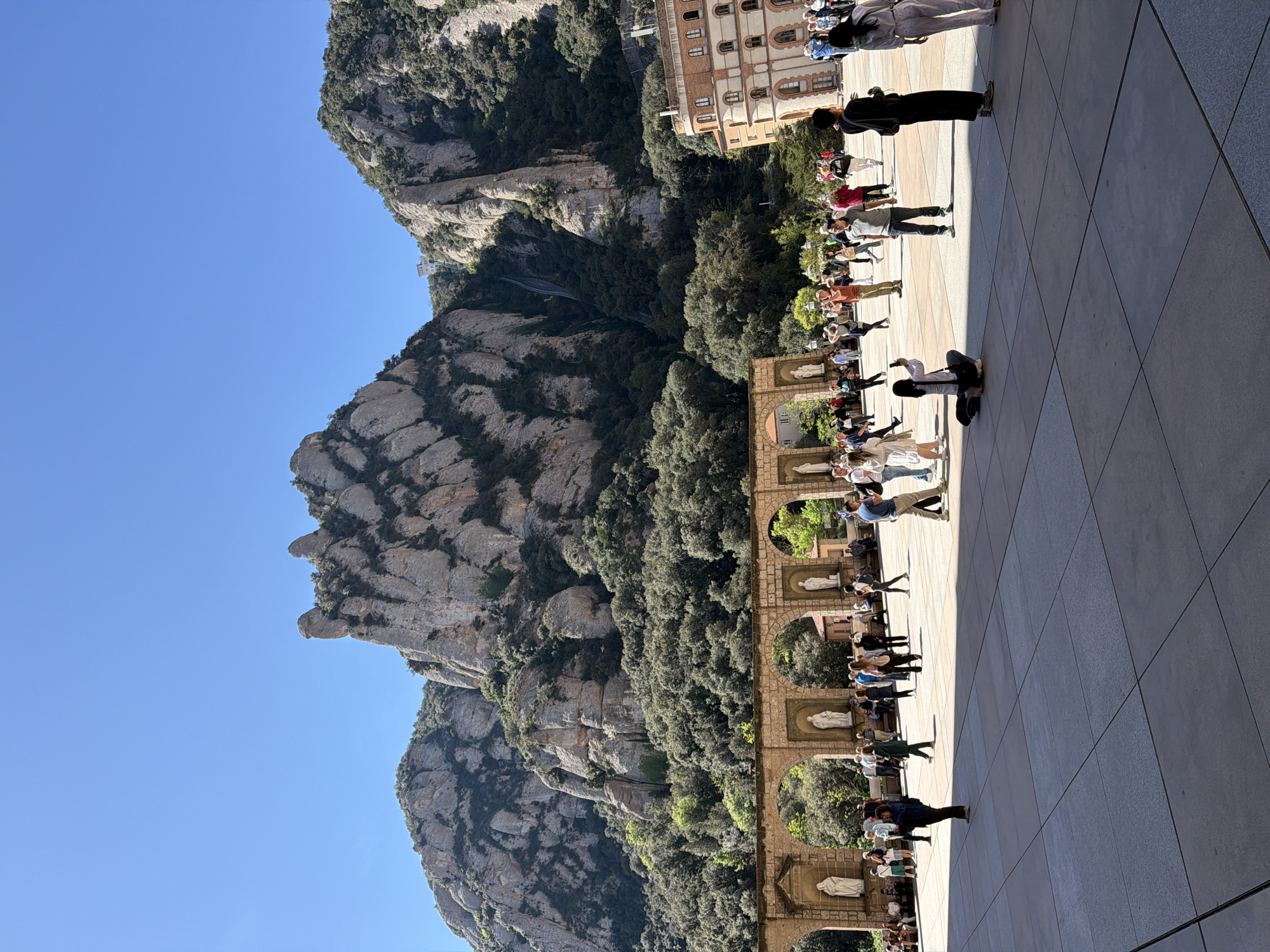 This image captures a bright, sunny day at the Montserrat complex in Monistrol de Montserrat, Spain, showcasing a blend of natural beauty and architectural heritage.
The scene is dominated by the majestic, rugged mountains of Montserrat, visible on the left and extending into the background. These distinctive rocky formations are partially covered in lush green vegetation. A clear, vibrant blue sky fills the upper portion of the frame. A faint funicular track can be observed ascending one of the mountain slopes in the distance, providing a key location cue.
In the mid-ground, a grand architectural structure stands on the right. This multi-storied building features a warm-toned facade with numerous arched windows. Below it, a series of ornate stone arcades stretches horizontally, each arch housing a white statue, likely depicting religious figures.
The foreground and right side of the image are occupied by a spacious, light-colored paved plaza. Numerous people, comprising adults and possibly some children, are scattered across this area. Many appear to be tourists or visitors, walking casually, standing and observing their surroundings, or gathering in what resembles a queue along the right edge of the plaza, partially shaded by the building.
The bright sunlight casts distinct, elongated shadows across the plaza, indicating a mid-morning or early afternoon time of day. The weather is clear and pleasant. No discernible text is visible anywhere in the image.
