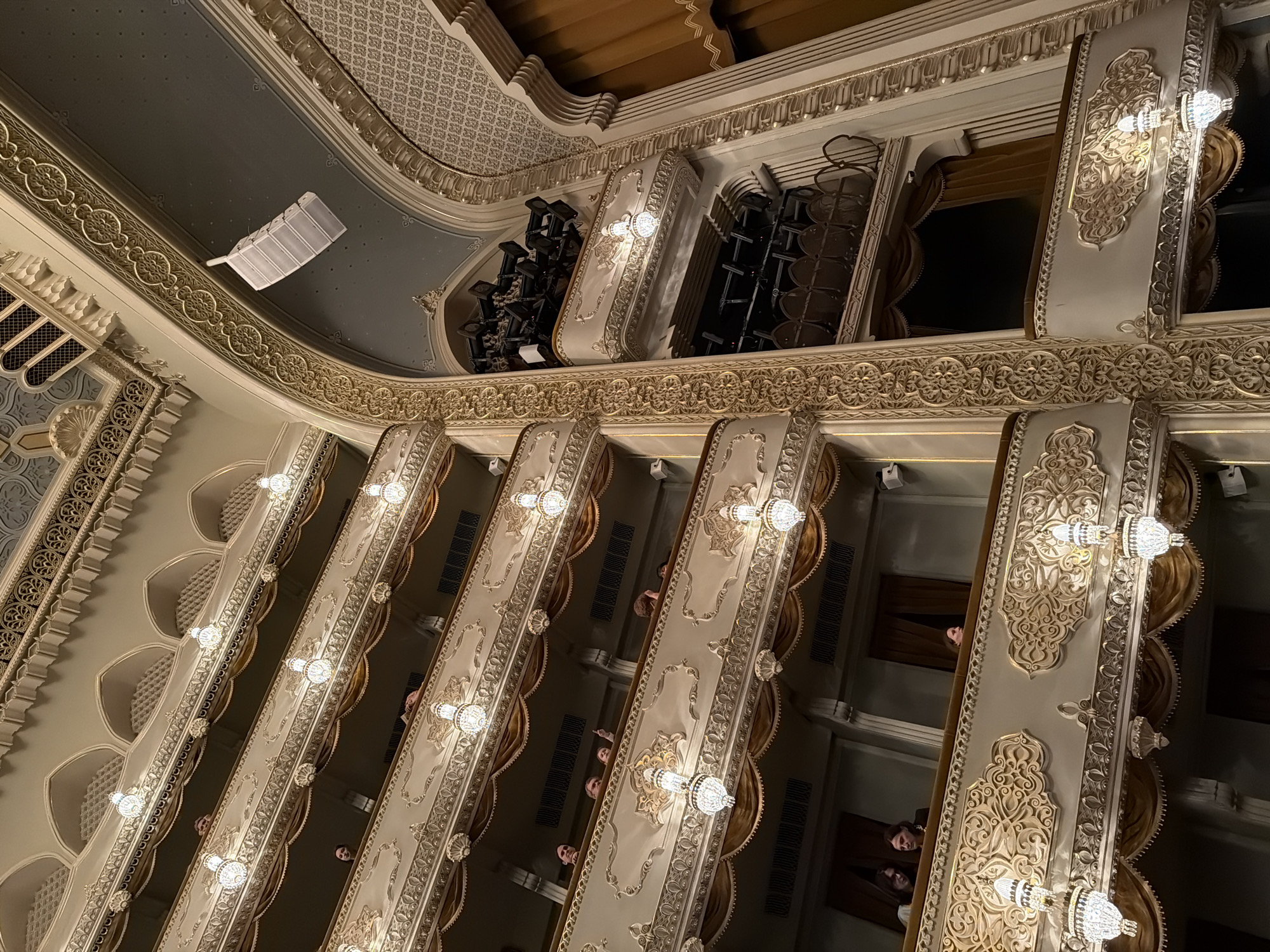 Captured from a lower vantage point, the image reveals the ornate, tiered balcony structure of the Tbilisi Opera and Ballet Theatre, a cultural landmark in Tbilisi, Georgia. The balconies, resplendent in shades of white and gold, are adorned with intricate carvings and lit by decorative sconces. A cluster of stage lighting equipment is visible on one of the upper tiers. Peering over the balconies, the faces of theatergoers can be discerned, adding a human element to the grand architectural scene. The ceiling of the theater is decorated with a geometric pattern.