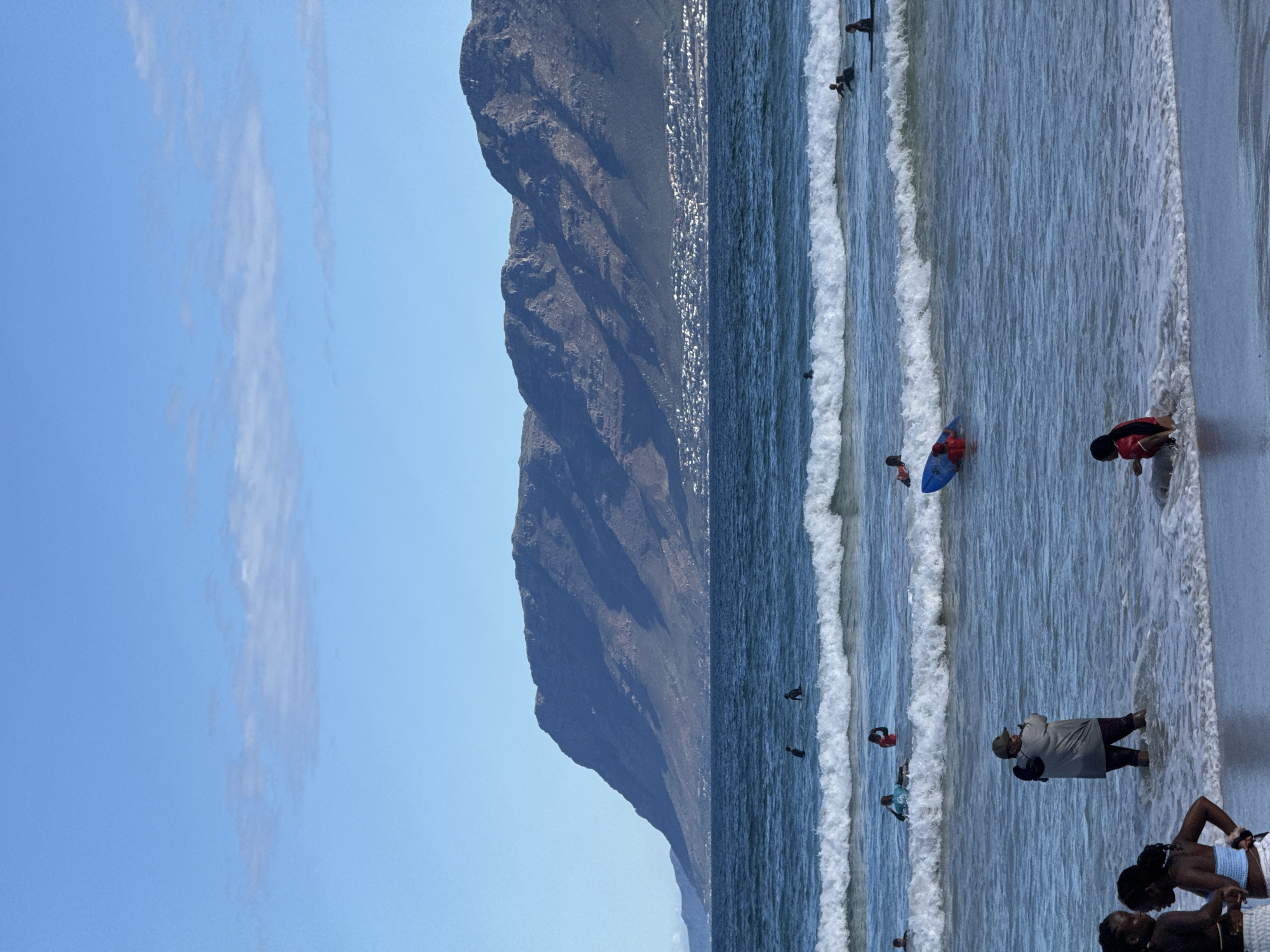 This is a medium shot of a beach scene in Cape Town, South Africa. Dominating the left side of the image is a large, dark mountain. The sky is blue with some sparse, thin clouds. The beach itself is populated with people enjoying the water. Some are standing near the shoreline, while others are further out, either swimming or surfing on the waves. The water appears a dark shade of blue, reflecting the light of the sun. The waves are visible breaking near the beach. The image shows a typical sunny day at the beach, with a backdrop of a mountain landscape. The photo is rotated 90 degrees clockwise.