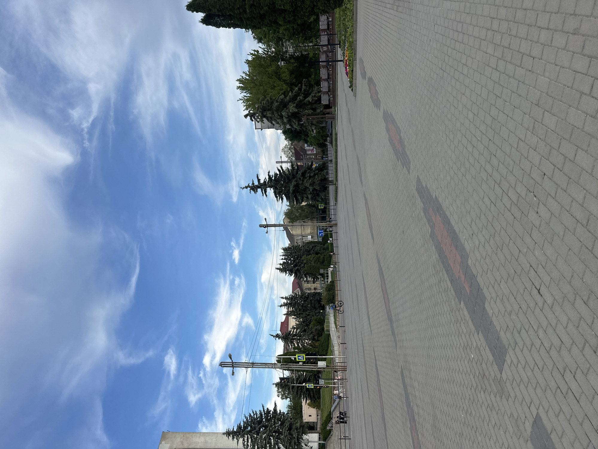 The image shows an outdoor plaza paved with grey interlocking bricks, featuring some red and dark grey rectangular inlays forming patterns. In the background, there are several evergreen trees, some deciduous trees, and buildings with red roofs. A tall, modern concrete building is visible on the left. A street with overhead power lines and traffic lights is present, along with street lamps. A person is riding a bicycle across the plaza, and a few other individuals and a stroller are visible near the background. The sky is blue with scattered white clouds. The scene appears to be a public square in the city of Ternopil, Ukraine.