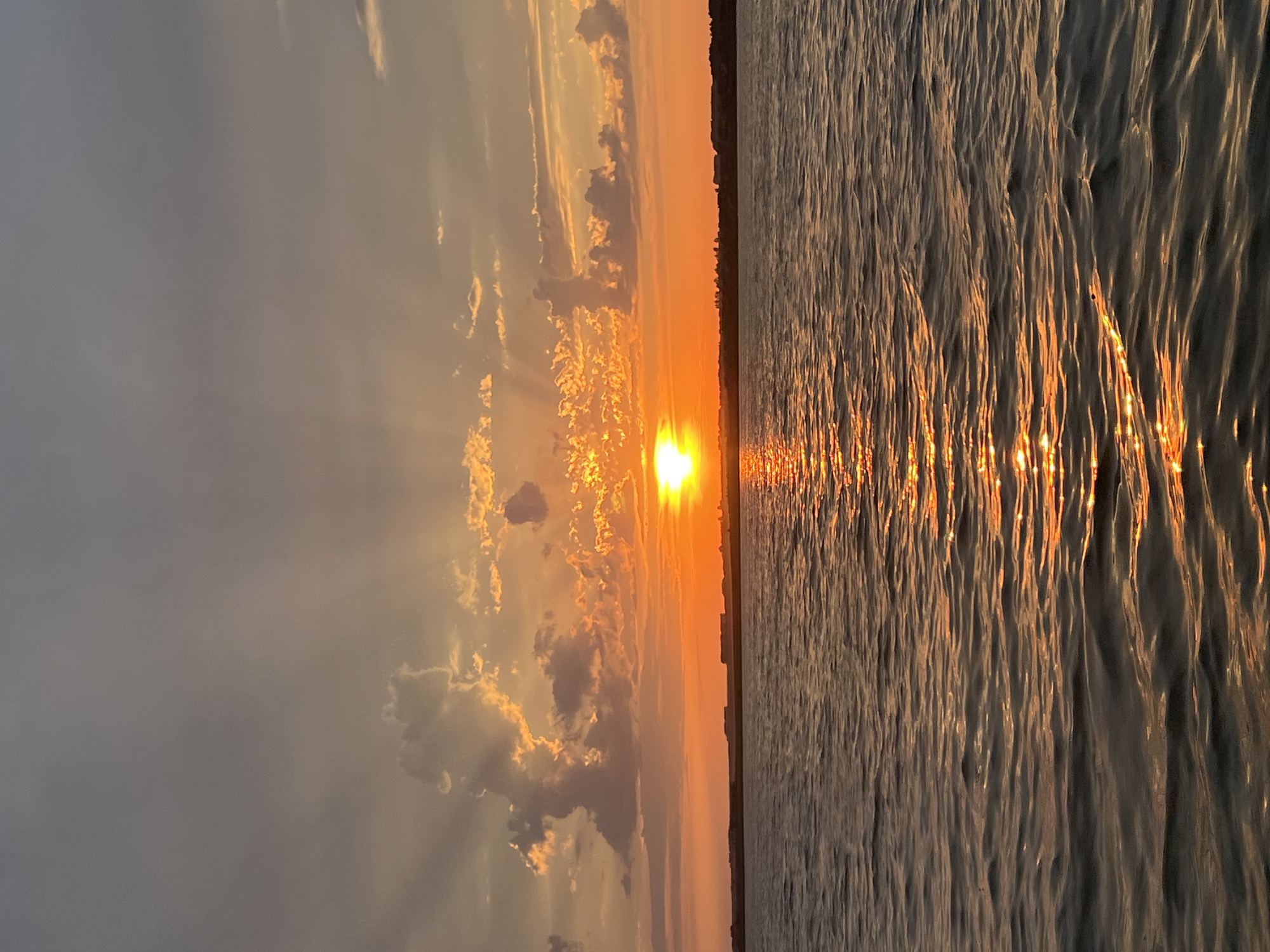 A tranquil body of water, likely a lake or large river, fills the foreground of the image. The water's surface is gently rippled, reflecting the warm hues of the sunset. In the mid-ground, a low, flat horizon line separates the water from the land. This land appears to be sparsely populated, with a few indistinct buildings or structures visible along the distant shore. The background is dominated by a dramatic sunset sky. The sun, a bright orange orb, is positioned just above the horizon, casting long, ethereal rays through scattered clouds. The clouds themselves are illuminated with vibrant shades of orange and yellow, creating a striking contrast against the cooler grey tones of the upper sky. The overall scene evokes a sense of peace and natural beauty, consistent with a sunset over a body of water in a relatively undeveloped area. The location is identified as Novopichugovo, Russia.