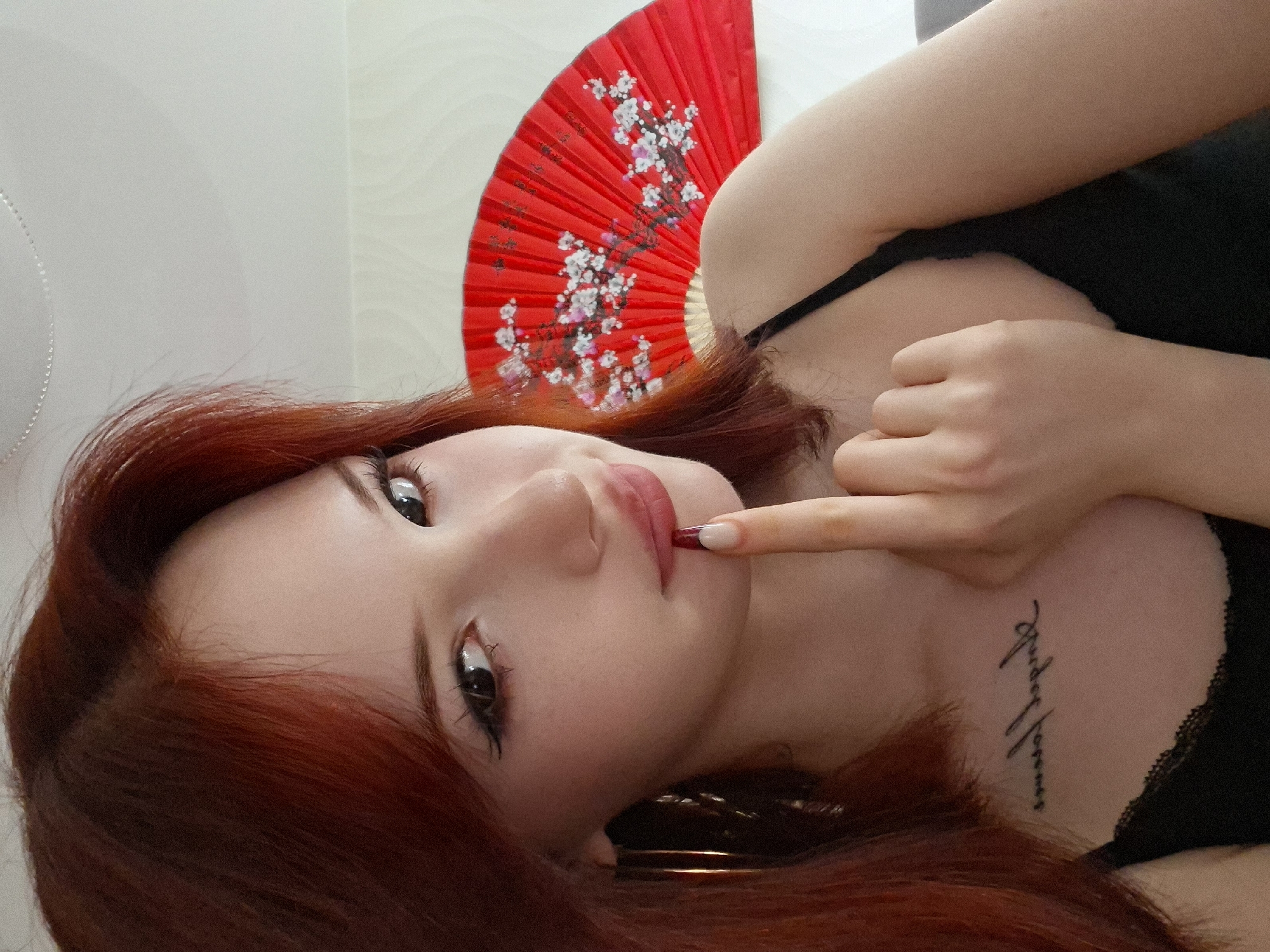 A young woman with auburn hair is reclining and facing the camera. She is wearing a black spaghetti strap top and has a tattoo on her upper chest. Her index finger is touching her lips, and she has makeup on. Behind her is a partially visible red hand fan with a white floral design. The background appears to be a light-colored wall. The image does not contain information about Chaikovsky, Russia.