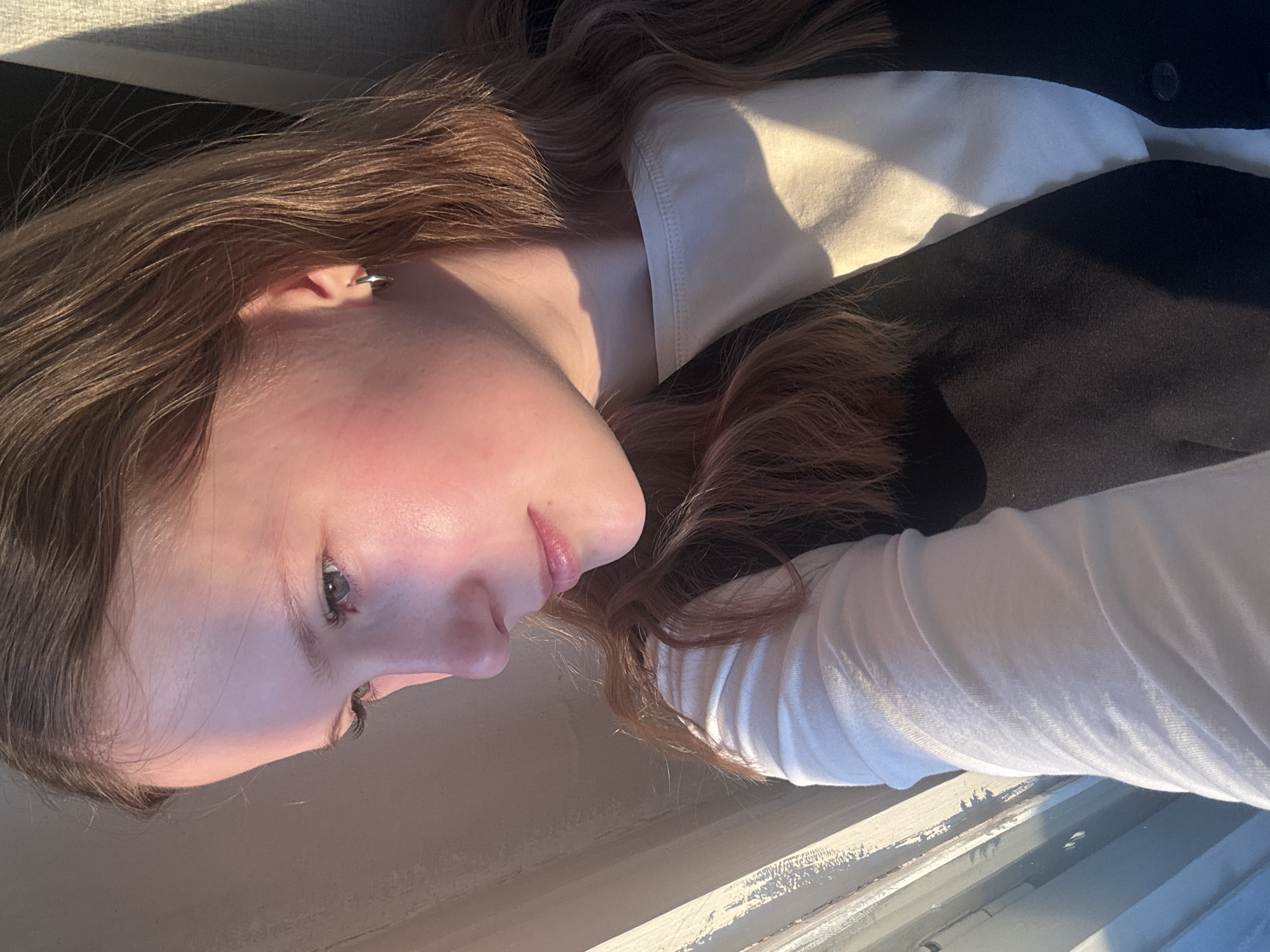 The image shows a close-up, tilted portrait of a young woman with light brown hair, fair skin, and blue eyes. She is wearing a white long-sleeved shirt underneath a dark vest or jacket. The light source appears to be sunlight from a window, casting shadows across her face and clothing. The background is indistinct, showing parts of the window frame.
The location is not discernable from the provided image and cannot be attributed to Chaikovsky, Russia. There is no information present to suggest a specific geographical location.