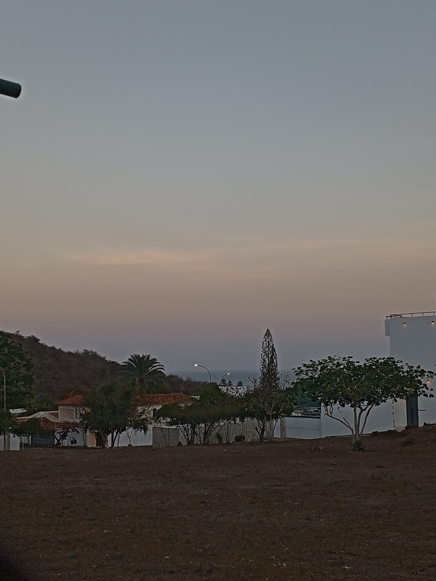 The image depicts a landscape at dusk in Pampatar, Venezuela. In the foreground, there is a field of dry, brown earth. Beyond the field, a row of trees and vegetation obscures parts of houses and buildings. Some houses have red tile roofs and white walls. In the middle ground, there are palm trees and other trees. The background shows a hill on the left side and a glimpse of the ocean towards the center. The sky is a gradient of pale blue and orange, indicating either sunrise or sunset. A street lamp is visible amongst the trees. On the right, a white building stands, possibly a multi-story structure, and the surrounding environment is visible.