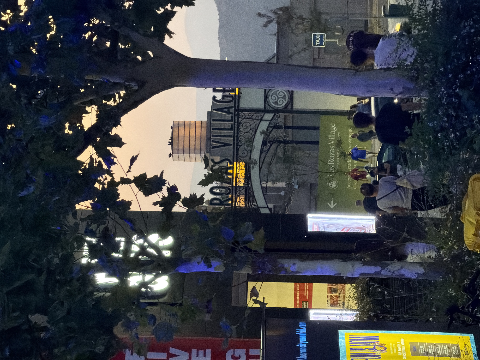 This is a nighttime photograph of the entrance to "Las Rozas Village," an outlet shopping center in Las Rozas de Madrid, Spain. The entrance archway features illuminated text that reads "LAS ROZAS VILLAGE." In the foreground, several trees with dark foliage are visible, some with blue lights reflecting on their trunks. The sky above is a muted orange hue, indicative of dusk or dawn.
Several people are visible in the scene. In the middle ground, a group of individuals are walking towards or through the archway. On the right side of the image, two individuals are standing, one wearing a white shirt with dark patterns and the other wearing a dark shirt. Further to the right, a "TAXI" sign is mounted on a pole.
Various signs are present, including a bright yellow sign on the left with text that appears to be advertising events. Another illuminated sign behind a tree reads "FIVE GUYS REWARD." A green sign with "Las Rozas Village" written on it is also visible. The environment appears to be an urban outdoor setting with paved ground and some greenery.