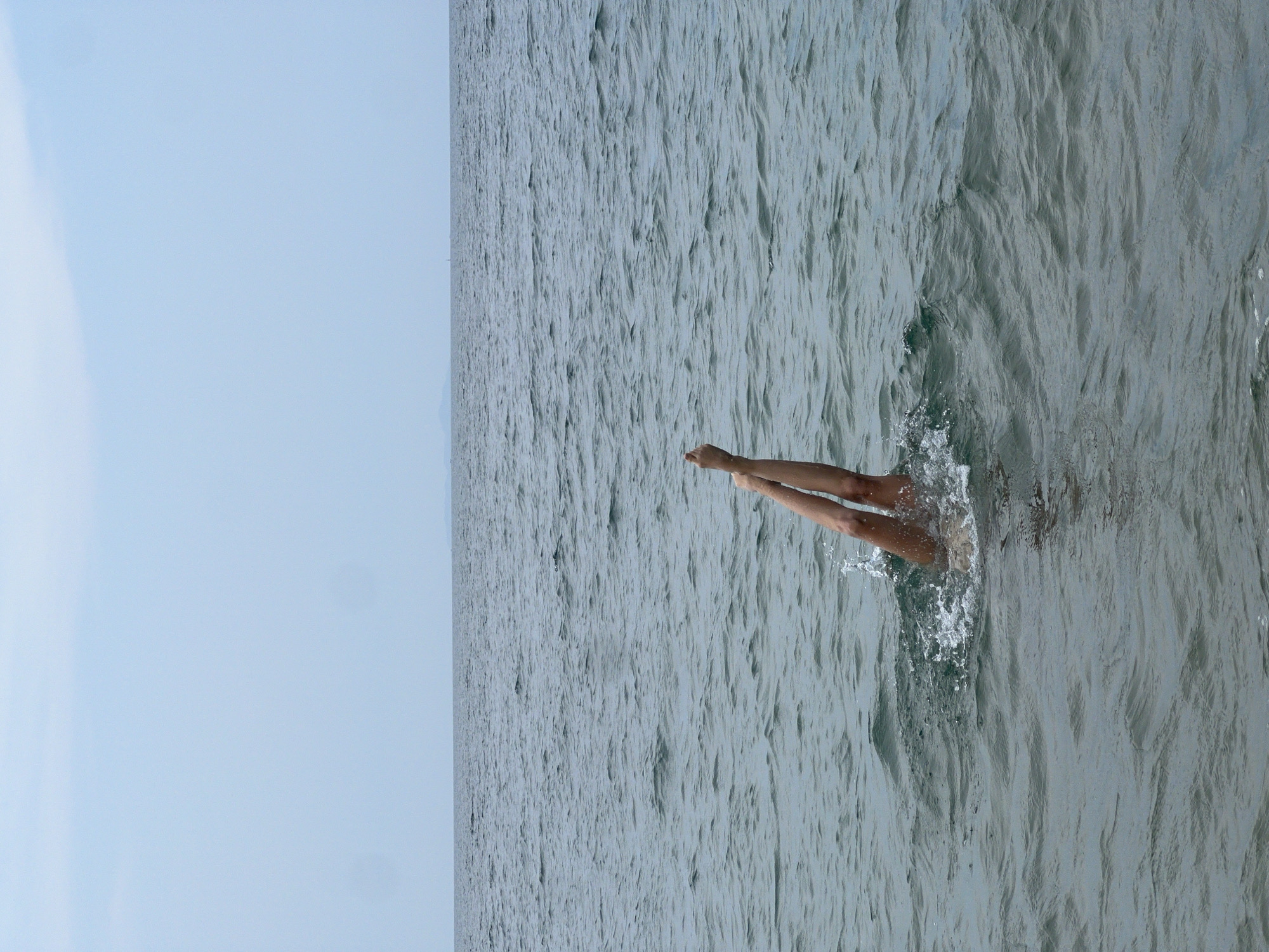 The image captures a dynamic moment in a body of water under a clear sky. A person's legs are visible, extending out of the water with splashes and ripples around them, suggesting an active movement like diving or jumping into the sea. The legs are a light skin tone, positioned almost vertically, with the feet pointed upwards.
The water surface, covering the majority of the image, is a muted grey-green, characterized by numerous small waves and undulating patterns. The immediate area around the legs shows white foamy splashes, indicating the force of entry or exit. The horizon line is visible, separating the water from a bright, light blue sky that occupies the upper left portion of the image. The sky is clear, with no clouds visible.
The scene conveys a sense of outdoor recreation on a bright day. The vastness of the water and sky indicates an open body of water, likely a sea or ocean. Given the provided context, this scene is set in Nha Trang Ward, Vietnam. No other subjects or visible text are present in the image.