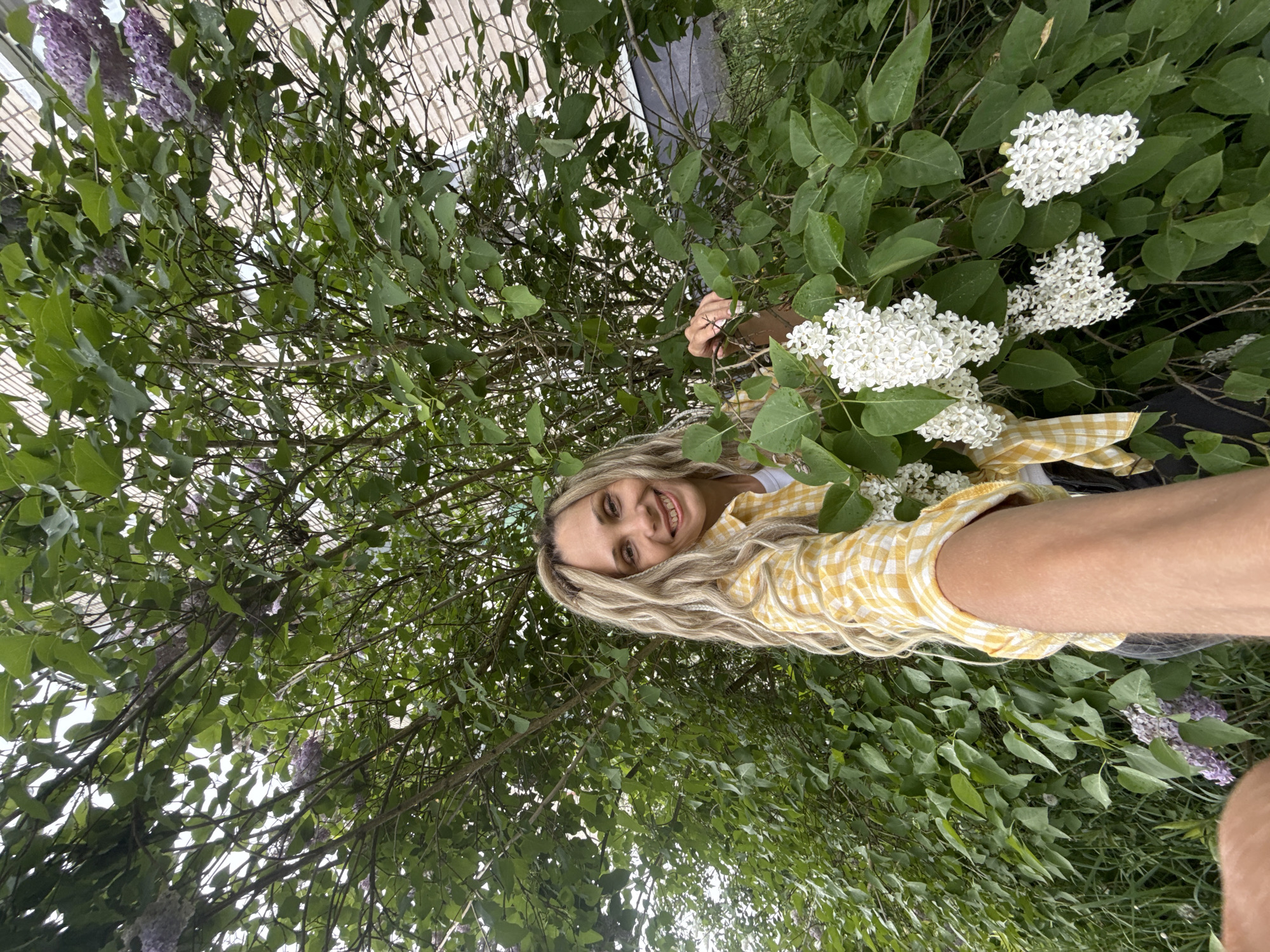 A woman with blonde hair, wearing a yellow and white checkered shirt, is smiling at the camera. She is surrounded by green foliage and white flowers, likely lilac bushes. The photo appears to be taken from a low angle, looking up at the woman and the trees. The location is assumed to be Rusanovo, Russia, though specific details within the image to confirm this are absent.
