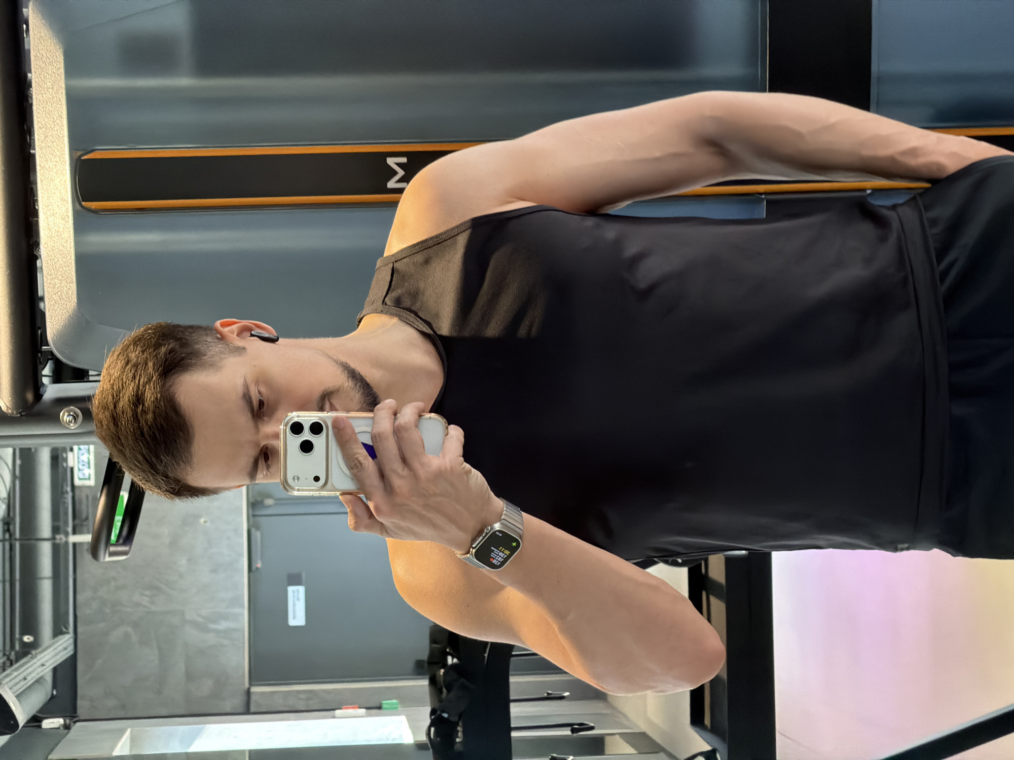 A male adult, appearing to be in his 20s or 30s, is captured in a mirror selfie within what seems to be a modern gym or fitness center, potentially in Krasnoyarsk, Russia. The man has short brown hair and a neatly trimmed beard, and is wearing a black sleeveless top (tank top) and black athletic shorts.
He holds a light-colored smartphone with a clear case in his right hand, angled to take the picture. A black earbud is visible in his left ear. On his left wrist, he wears a rectangular smartwatch with a metallic band. The watch face displays various digital information, including "17:36" (likely time) and a series of numbers which appear to be fitness metrics: "3,293", "2,661", "1,797", and "853".
The background features gym equipment, notably a piece with an orange stripe and a white 'Σ' (Sigma) symbol on a dark panel, set against a grey metallic surface. Other reflective surfaces and elements of exercise machines are also visible, reinforcing the gym setting. The lighting suggests an indoor, artificially lit environment. The man's expression is neutral and focused on taking the selfie.