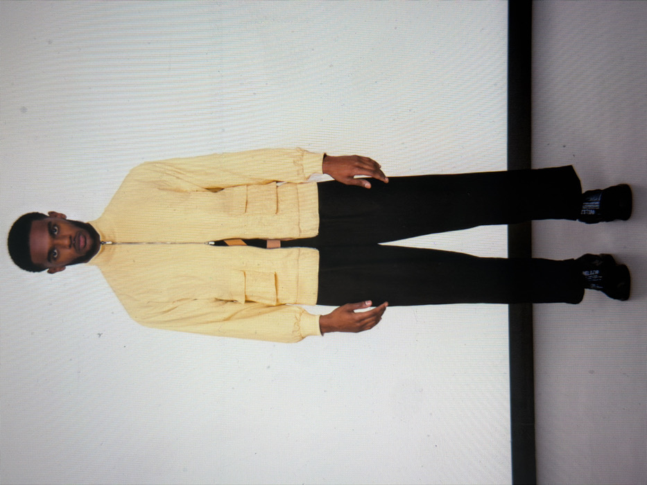 A male individual with dark skin and short, dark hair stands centrally, facing forward against a plain white background. He is wearing a light yellow, textured, long-sleeved jacket with a high collar and a silver zipper running down the front, partially open to reveal a dark garment with yellow or light-colored vertical stripes underneath. The jacket features two stacked, rectangular flap pockets on each side of the torso. He wears loose-fitting, wide-legged black trousers and dark athletic shoes with white text or a pattern visible on the outer sides. His hands hang relaxed at his sides, just below the jacket sleeves. The environment is a studio setting with a clean, light-colored floor, possibly dark grey, delineated by a distinct black horizontal line where it meets the white wall. No specific location within the city of London, United Kingdom, can be identified from the visual information presented.