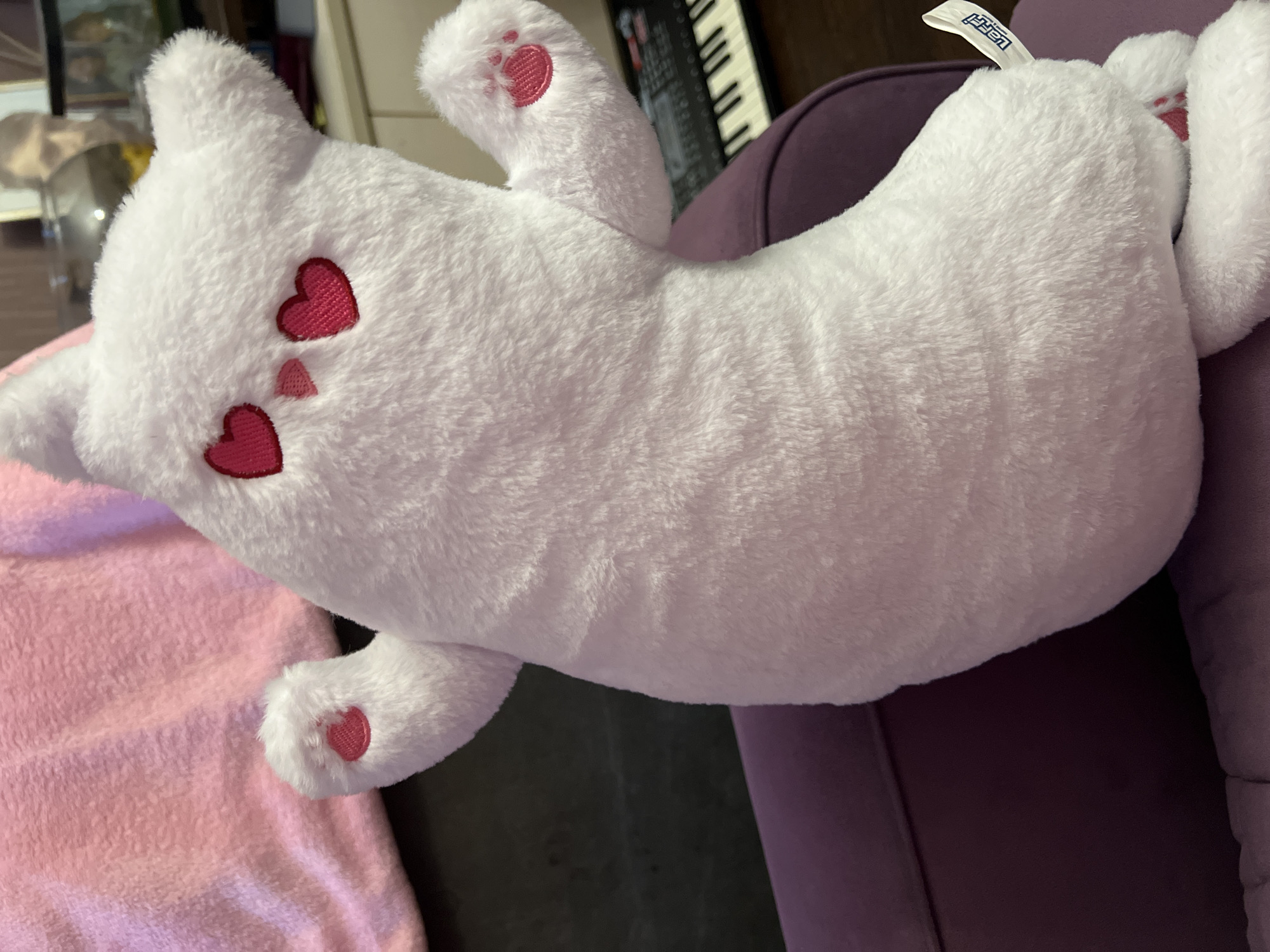 A white, plush cat toy is lying on a purple chair with a pink blanket nearby. The toy has two red heart shapes stitched on its face and a small red paw print on one of its feet. A keyboard is visible in the background. The scene appears to be indoors.