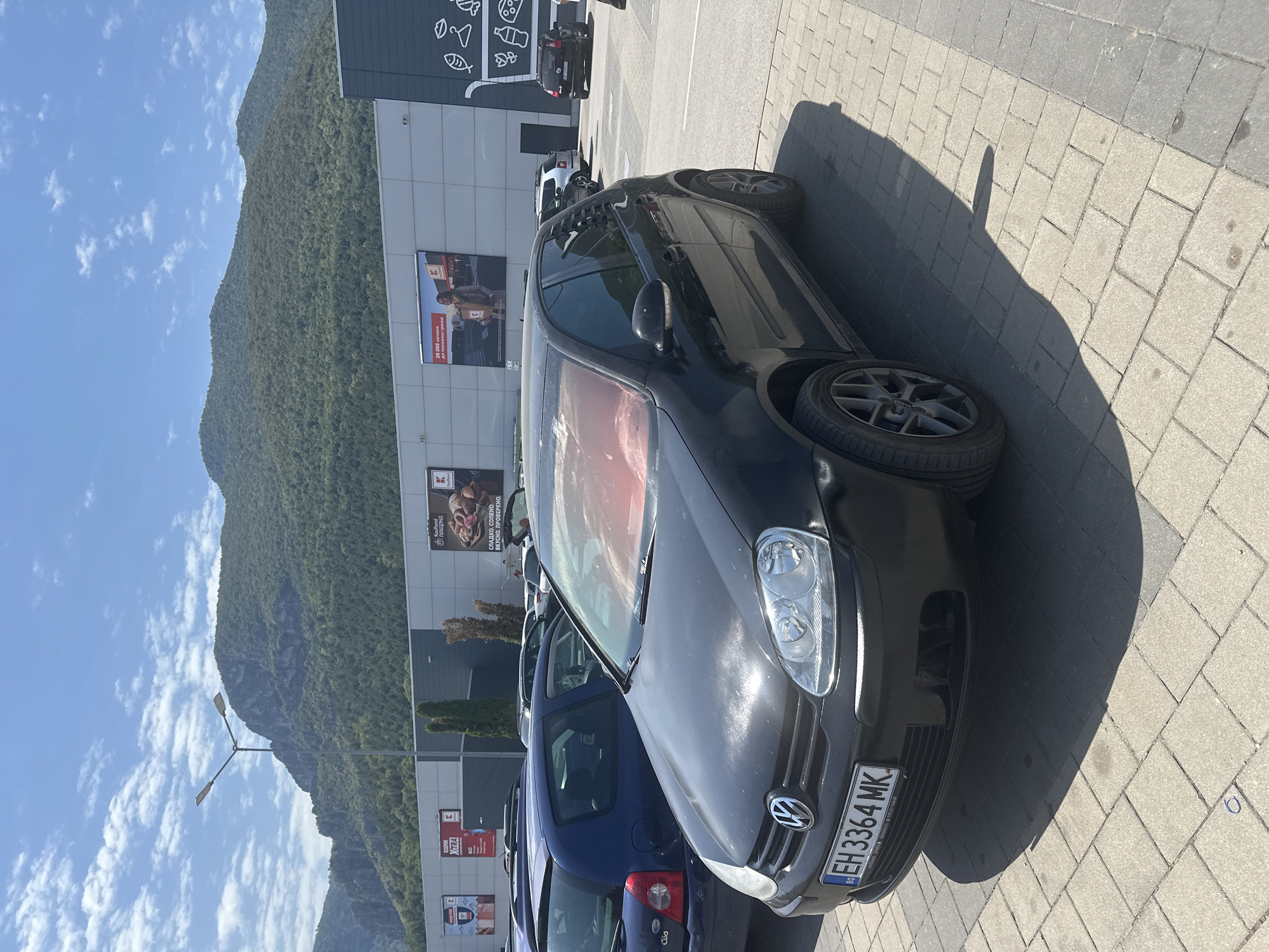 In a parking lot located in the city of Vratsa, Bulgaria, a black Volkswagen Golf is parked in front of a large retail building. The car, bearing the license plate EH 3364 MK, has its front end facing the viewer. The windshield appears to have a red tint from what may be sunshades or an overlay. Behind the car, other vehicles are visible, including a blue hatchback. The retail building has several large advertisements displayed on its facade, with one featuring a woman holding a box and another showing baked goods. The background consists of a steep, tree-covered mountainside under a partly cloudy blue sky, suggesting an outdoor commercial area at the edge of a mountainous region. The parking lot is paved with interlocking concrete blocks.