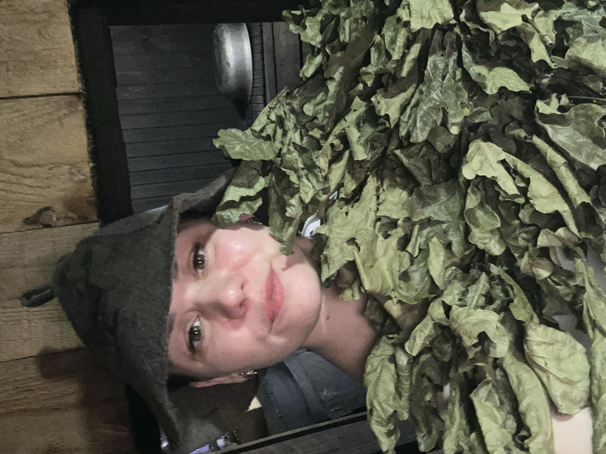 A person wearing a felt hat and covered with dried leaves is in what appears to be a wooden sauna. The location may be in Chaikovsky, Russia. The individual is turned slightly towards the camera and appears to be posing. The sauna has wooden walls and a dark interior. A metallic object is visible on the wall behind the person's head. The image is somewhat dark and grainy.