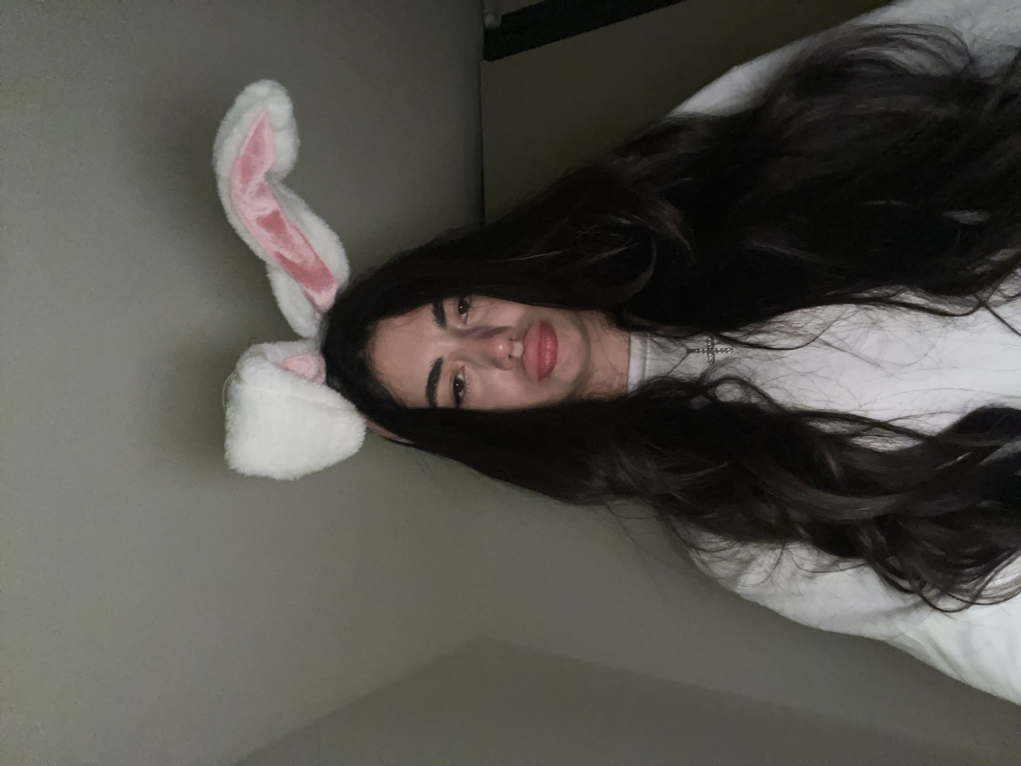 A young woman with long, dark hair and bunny ears on a headband is looking at the camera with her mouth slightly pursed. She is wearing a white sweatshirt and a silver cross necklace. The background appears to be an indoor setting with a plain wall. The image was taken on June 1, 2023, at 11:46 PM. No specific location in Chotěšov, Czechia is provided in the image data.
