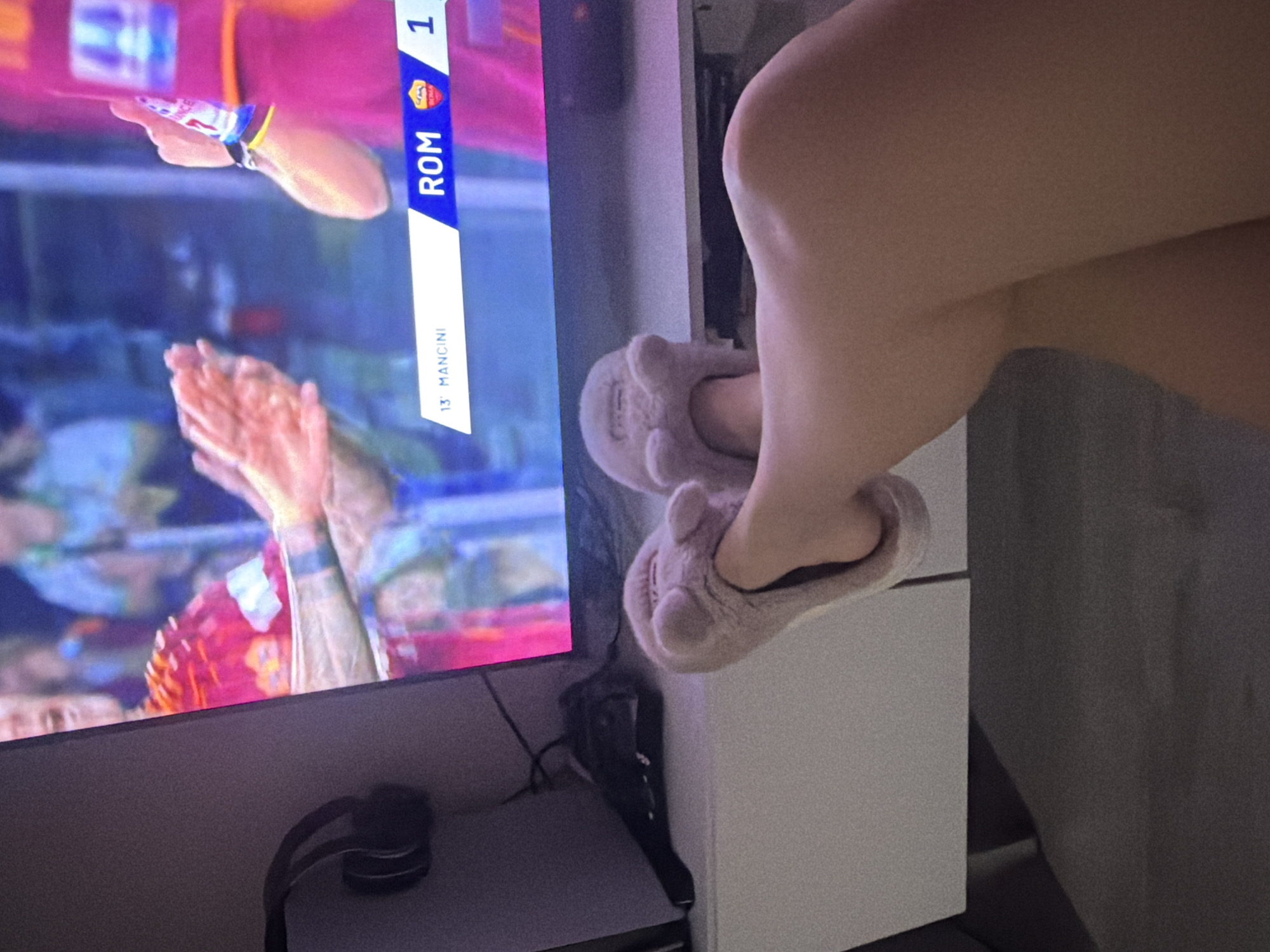 A television displays a soccer game with the score ROM 1. Headphones are located on a white shelf below the television. A person wearing fuzzy slippers is seated next to the television. The location is unknown.