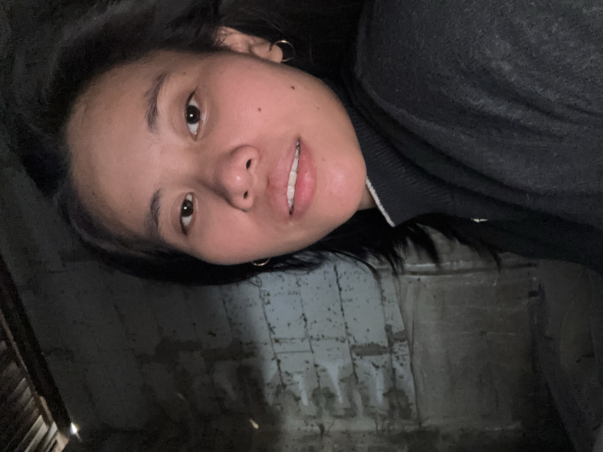 A woman is taking a selfie in what appears to be a dimly lit, unfinished room. The background is a rough brick wall with some discolored patches and what looks like wooden beams or roofing material at the top. The woman is wearing a dark sweater with a white trim around the collar. She has dark hair and is wearing small hoop earrings. Her facial expression is neutral, and she is looking directly at the camera. The lighting is low, casting shadows on her face and in the room.