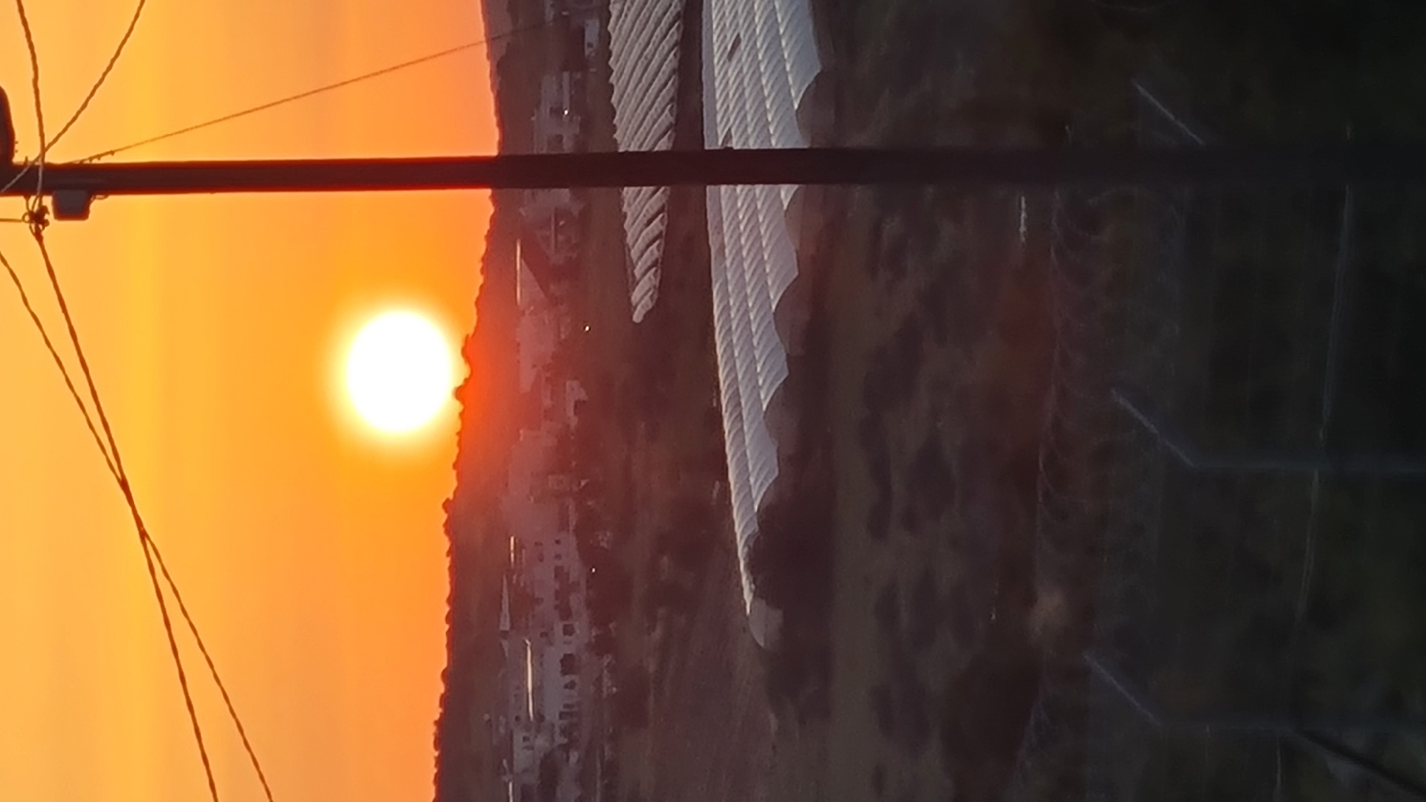 The image captures a sunset in what appears to be a rural or semi-rural area within the Lachish Regional Council, Israel. The sun is a bright orb in the orange sky. In the foreground, there are utility poles and wires, suggesting the presence of infrastructure. A cluster of buildings is visible, situated on a slope. The background features what seem to be long, low-rise structures, possibly greenhouses, amid a field.