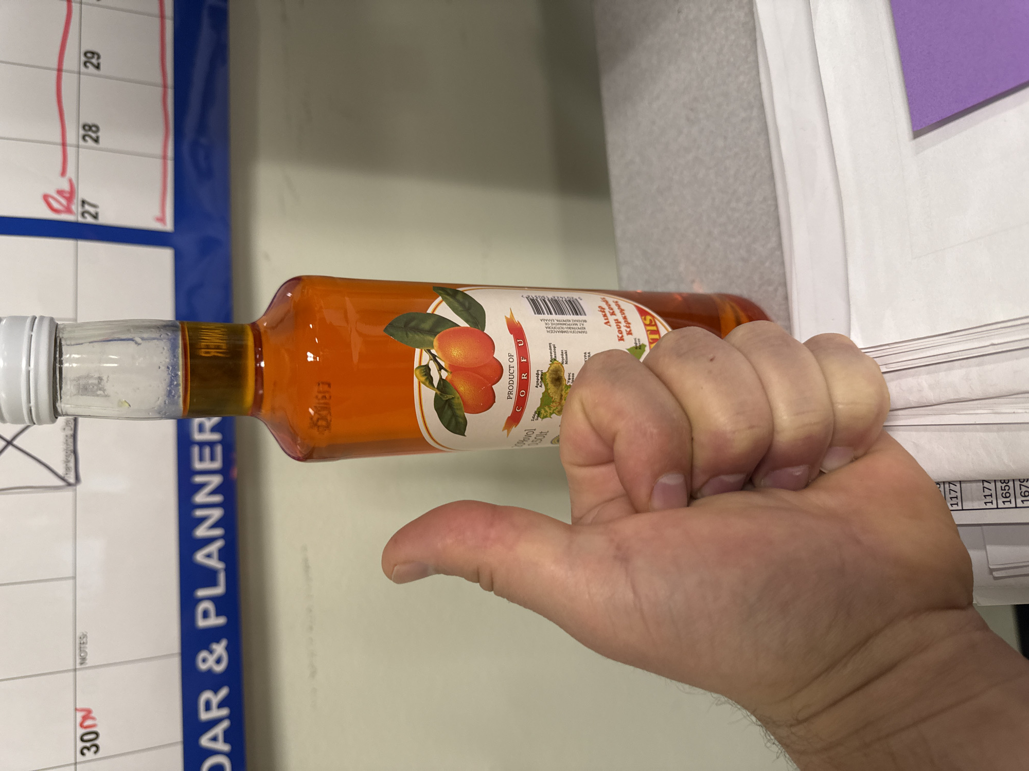 The image shows a person's thumb giving a thumbs-up gesture in front of a bottle of orange-colored liquid. The bottle has a label with images of fruit and text in English and Greek. Behind the bottle and thumb, a portion of a calendar is visible, with dates marked for November. The overall environment appears to be an indoor setting, possibly an office or a home, with a desk or table in the foreground. There are no discernible landmarks that would place this image specifically in the city of Kent, United States of America.