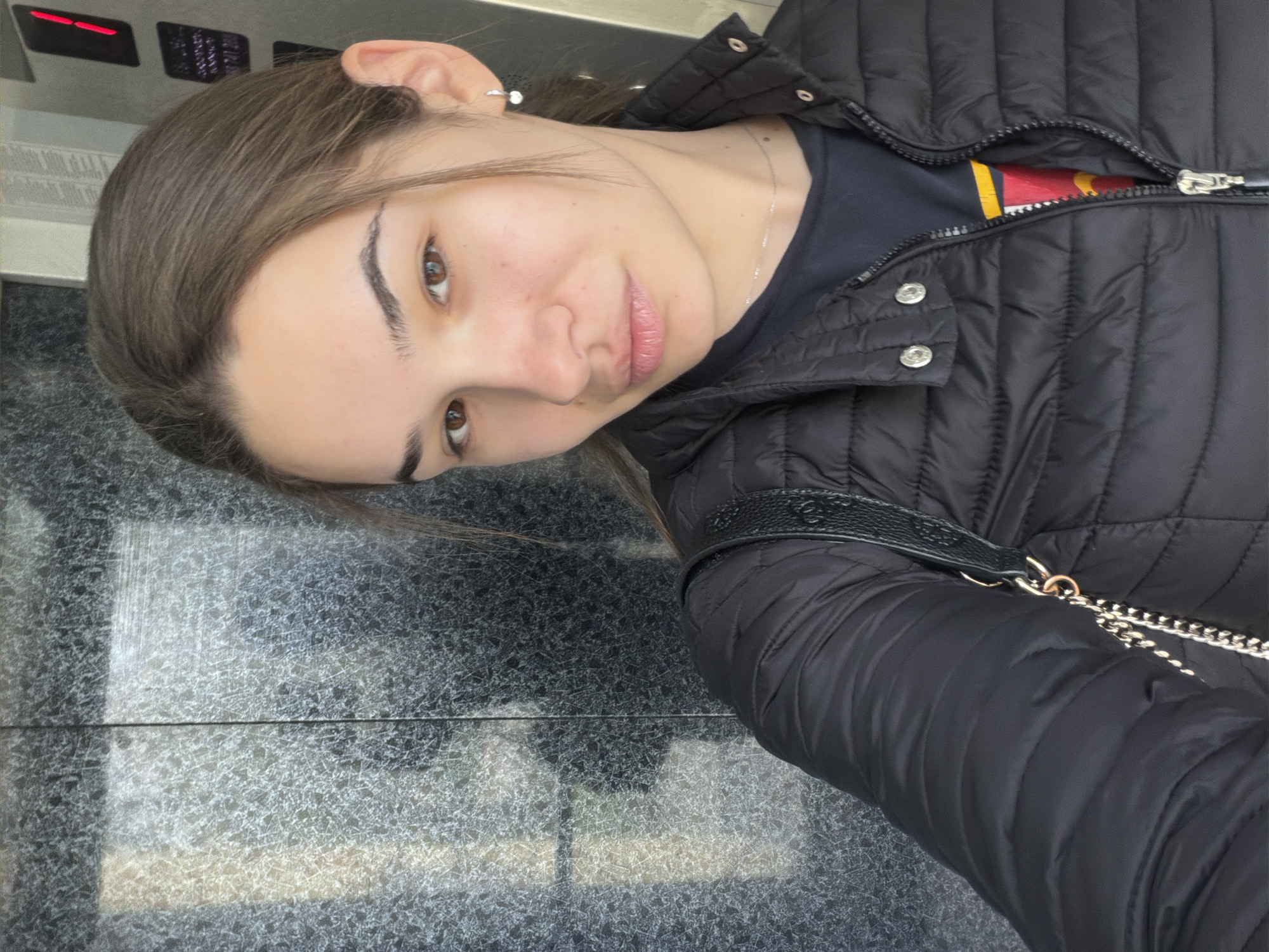 A young woman is in what appears to be an elevator. The walls of the elevator are made of polished gray marble. She is wearing a black puffer jacket, unzipped to reveal a dark t-shirt and a gold necklace. A black leather purse with a silver chain is visible on her shoulder. The woman is facing the camera with her head tilted to the side. Her hair is light brown, and she has fair skin and brown eyes. In the upper left corner, there is an electronic panel with red and black displays. It is not possible to determine that the location is in Sofia, Bulgaria.