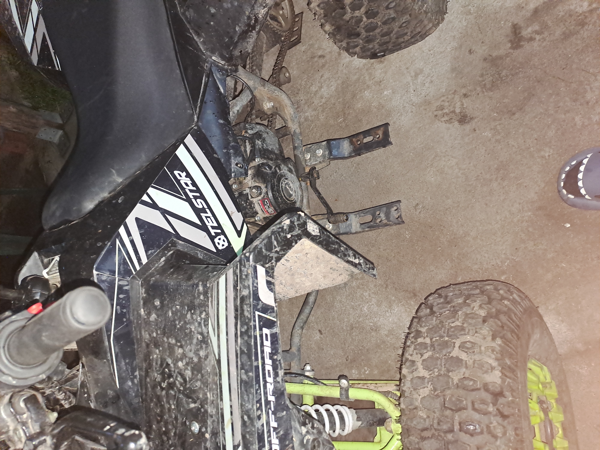 The image depicts a partially visible Telstar ATV in Obruchishte, Bulgaria. The ATV is dirty, with visible mud on the body and tires. The front left portion of the ATV is in focus, showing the handlebar, seat, engine, and front tire. The vehicle is parked on a concrete surface indoors. A single shoe is visible on the concrete floor near the right side of the image. The overall lighting is dim, and no people are visible in the frame.