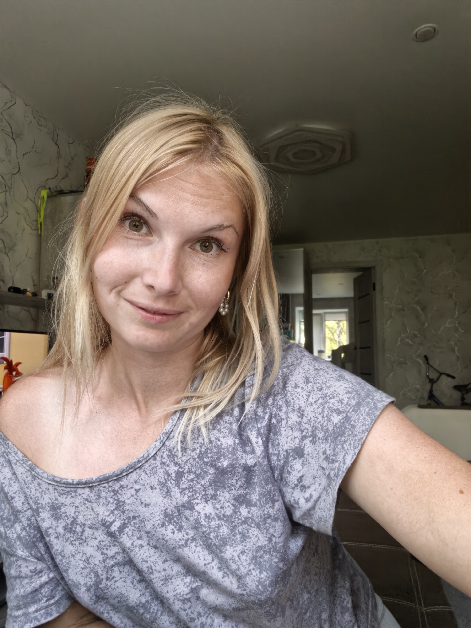 A young Caucasian woman with blonde hair and light skin is pictured from the chest up, looking directly at the camera. She is wearing a grey, mottled t-shirt that is off-the-shoulder. Her expression is a slight smile, and her eyes are brown. She has freckles on her cheeks and nose, and is wearing pearl earrings. The background shows an indoor setting with textured walls and a ceiling with a decorative fixture. A doorway leading to another room is visible, with a glimpse of trees and sunlight through a window. The image quality is fair, suggesting it was taken with a standard camera or smartphone. The location is described as Sterlitamak, Russia, however, there are no visual elements in the image that specifically identify this location.