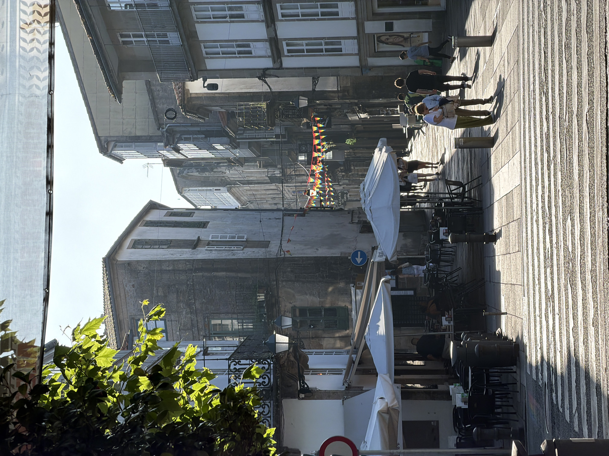 The image depicts a street scene in Santiago De Compostela, Spain, likely in the historic old town, bathed in the warm light of late afternoon. The perspective is from a pedestrian crossing, looking down a narrow street lined with multi-story stone buildings.
In the foreground, a zebra crossing is prominently featured, with harsh shadows cast across it, indicating the low angle of the sun. To the left, several white patio umbrellas shade tables and chairs belonging to an outdoor cafe. A large tree with vibrant green leaves is visible in the upper left corner, its foliage backlit by the sun, creating a dappled effect.
Moving further down the street, people are seen walking and interacting. On the right side, a couple is engaged in conversation near a bollard. Further down the street, several individuals are walking away from the camera. On the left, a woman in a light-colored top and white skirt walks away from the camera, and another person, mostly obscured, is also walking in that direction. A man in a dark shirt and shorts is walking towards the right side of the frame, and another person in a dark top and grey pants is walking away from the camera towards the right.
The buildings lining the street exhibit traditional Galician architecture, with stone facades, balconies, and numerous windows, some with shutters. Decorative banners, possibly for a festival, are strung across the street in the middle ground, adding a touch of color. The street is paved with cobblestones and rectangular stone slabs, typical of older European cities. The overall atmosphere is one of a bustling, sunny day in a historic urban environment.