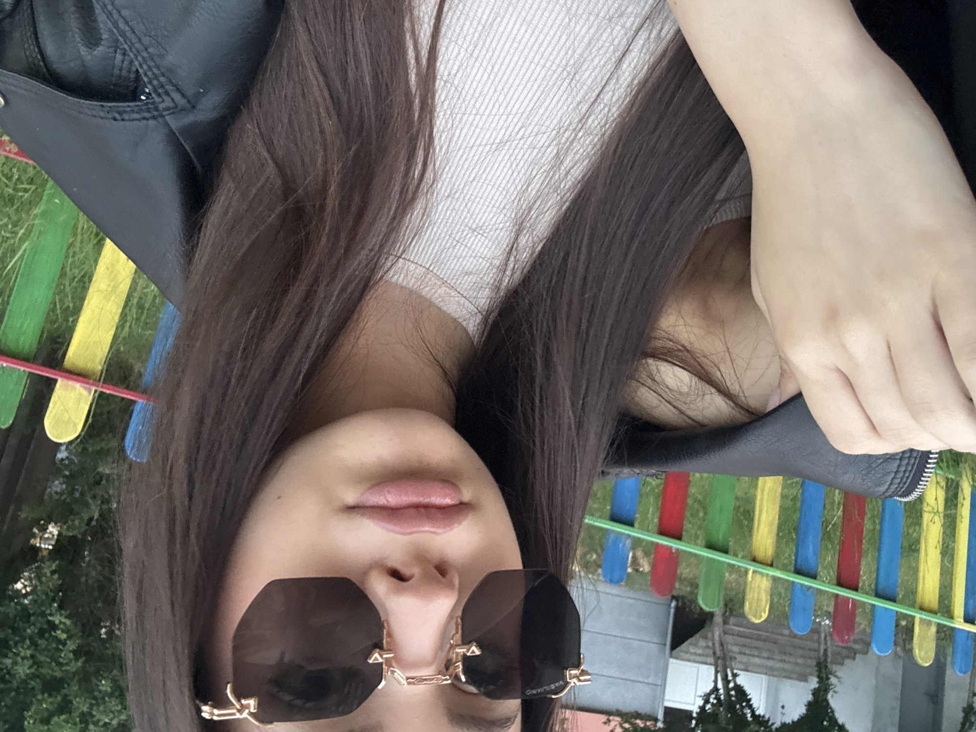 A close-up, chest-up outdoor portrait captures a person with long, straight dark brown hair, obscured by dark hexagonal sunglasses with gold-colored frames. The left sunglass frame (viewer's perspective) displays the text "VASILLIARKO" in gold. The person wears a light-colored, possibly off-white or light beige, ribbed top and a black leather-like jacket with a prominent zipper and collar detail on the right side. The left hand, with pale skin, is visible near the left shoulder, partially hidden by the jacket. In the background, out of focus, is a colorful fence composed of vertical slats in alternating blue, yellow, green, and red, with a horizontal green bar running across them. Beyond the fence, green grass, foliage, and parts of two buildings—one light-colored with visible stairs and another grey with a white door—are discernible. The setting is an outdoor area in the city of Unknown, Unknown.