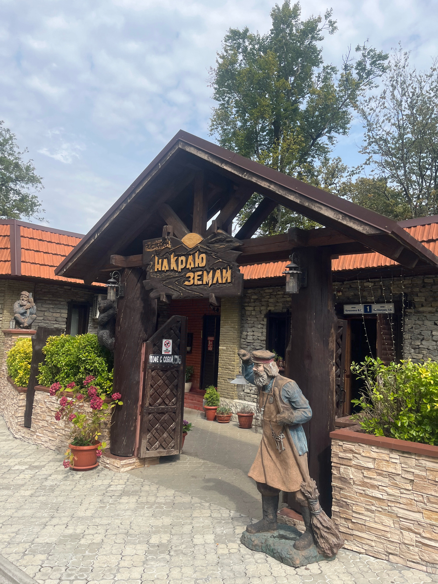 The image shows the entrance to a restaurant called "Накраю Земли" in Khartsyz Pervyi, Russia. The entrance is made of wood and stone, with a large wooden sign above the doorway. To the left of the entrance, there is a statue of a man sitting on a ledge, and to the right, there is a statue of a man with a broom. The restaurant has a stone facade and a tiled roof. There are plants and flowers around the entrance, and the ground is paved with cobblestones. The sky is cloudy.