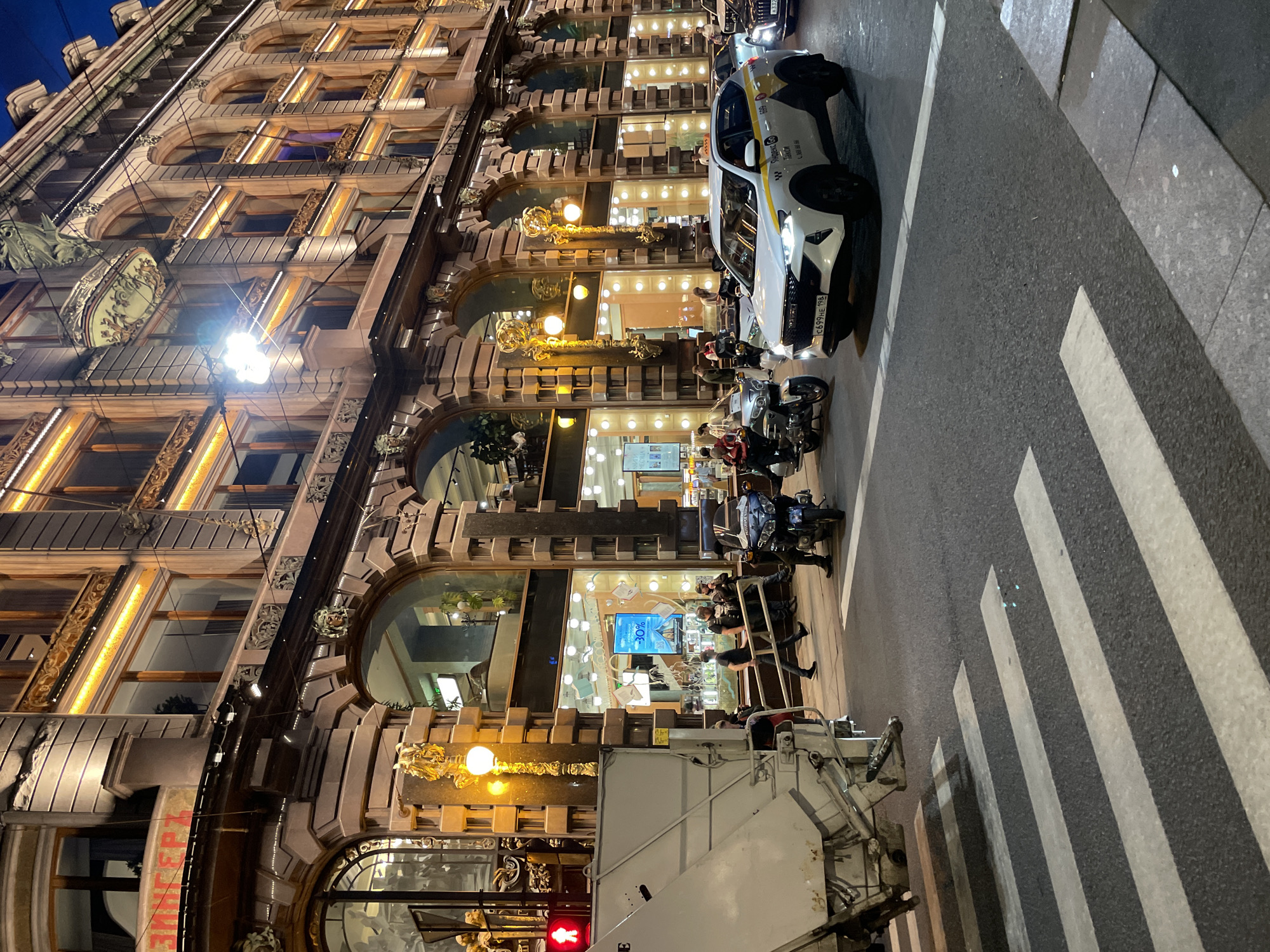 This image captures an evening street scene in front of the Singer House (Dom Knigi) located at Nevsky Prospekt, 28, Saint Petersburg, Russia. The prominent, multi-story building showcases ornate Art Nouveau architecture with numerous large arched windows on its lower levels, functioning as brightly lit display windows for a store. The facade is richly decorated with classical elements, including pilasters, cornices, and mascarons, all accentuated by warm yellow illumination tracing window frames and architectural features. A red sign displaying "ЗИНГЕРЪ" is visible on the upper left section of the building.
In the foreground, an asphalt road features a pedestrian crosswalk marked with white stripes. A white and yellow Yandex Taxi SUV, with license plate C699HE 198, is positioned in the middle-right of the road, facing slightly left. Two motorcycles are visible: a blue one with a seated rider on the right, and a silver one partially obscured by the taxi on the left. A dark sedan is partially visible behind the white taxi. On the far left, a large, light-colored refuse truck is parked, partially obscuring the sidewalk and a pedestrian signal displaying a red "standing man" symbol.
Several pedestrians are present on the sidewalk in front of the building and near the vehicles. A group of men stands near the blue motorcycle, and another man walks across the mid-ground. Inside the storefronts, various items of merchandise and a digital display advertising "30%" are visible. Ornate, golden-accented streetlights with multiple globes are positioned along the building's facade at street level. The scene is illuminated by a combination of the building's extensive lighting, streetlights, and vehicle lights, creating a vibrant urban night environment.