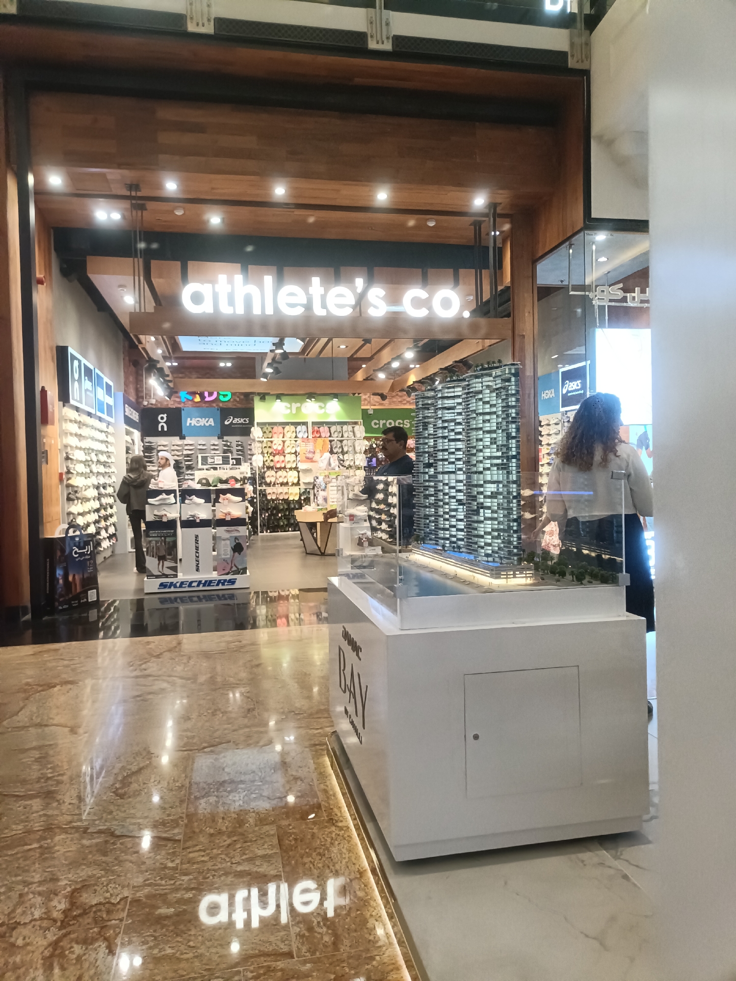 The image shows the entrance to an "athlete's co." store in Dubai, United Arab Emirates. The store is decorated with wooden panels and features large illuminated signs. Inside, shelves are stocked with shoes, and display stands showcase various brands like Skechers, Hoka, and Asics. A miniature architectural model of a building is prominently displayed on a white podium in the foreground. Two individuals are visible in the store: one is a customer browsing shoes, and another is a staff member assisting them. A third individual, a woman with curly hair, is seen from behind near a glass display. The floor is a polished marble, reflecting the store's lights. The overall environment suggests a modern retail space within a shopping mall.