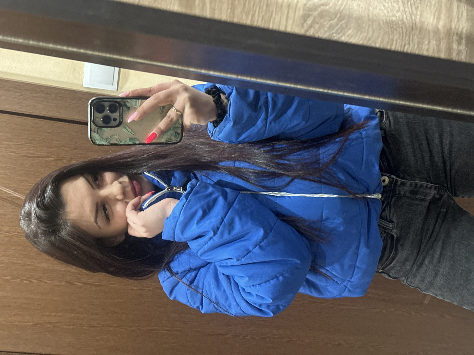A young woman with long brown hair is taking a selfie in a mirror. She is wearing a bright blue puffer jacket and dark jeans. The mirror is reflecting a portion of a room, including a wooden door and a light switch. The phone she is holding has a patterned case. The image does not depict the city of Sliven, Bulgaria.
