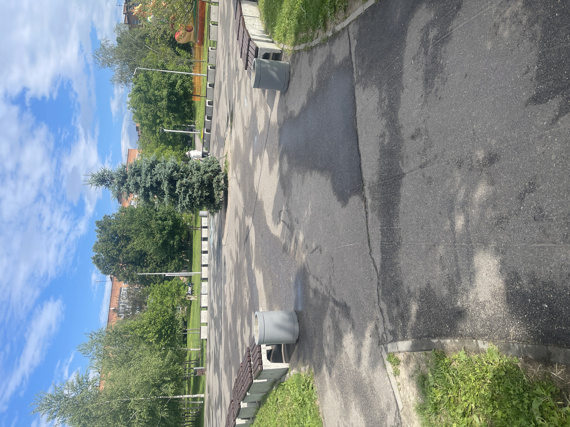 This media file depicts an outdoor urban park or public space in Poltava, Ukraine, on a bright, sunny day with a blue sky dotted with scattered white clouds. The image is oriented sideways, but when viewed correctly, it shows a wide asphalt pathway extending into the distance.
On the left side of the path, a prominent, large evergreen tree (likely a fir or spruce) stands amidst a variety of other deciduous trees, with lush green grass bordering the path. In the background, behind the trees, parts of brick buildings are visible.
The right side of the path features several public amenities: a series of concrete benches with wooden seats and a few cylindrical concrete bins or planters. The asphalt surface of the path shows variations in tone, with darker patches that could indicate moisture or wear, and is patterned with long shadows, suggesting the sun is not directly overhead, possibly mid-morning or late afternoon.
No specific activity or event is clearly depicted, implying a calm, everyday scene. While no text is directly visible, one of the provided hint crops reveals a large, stylized, reddish-brown object (possibly a character or animal head) peeking from behind trees in the far background, suggesting a children's play area or decorative park feature. The overall impression is that of a well-maintained, tranquil public area.