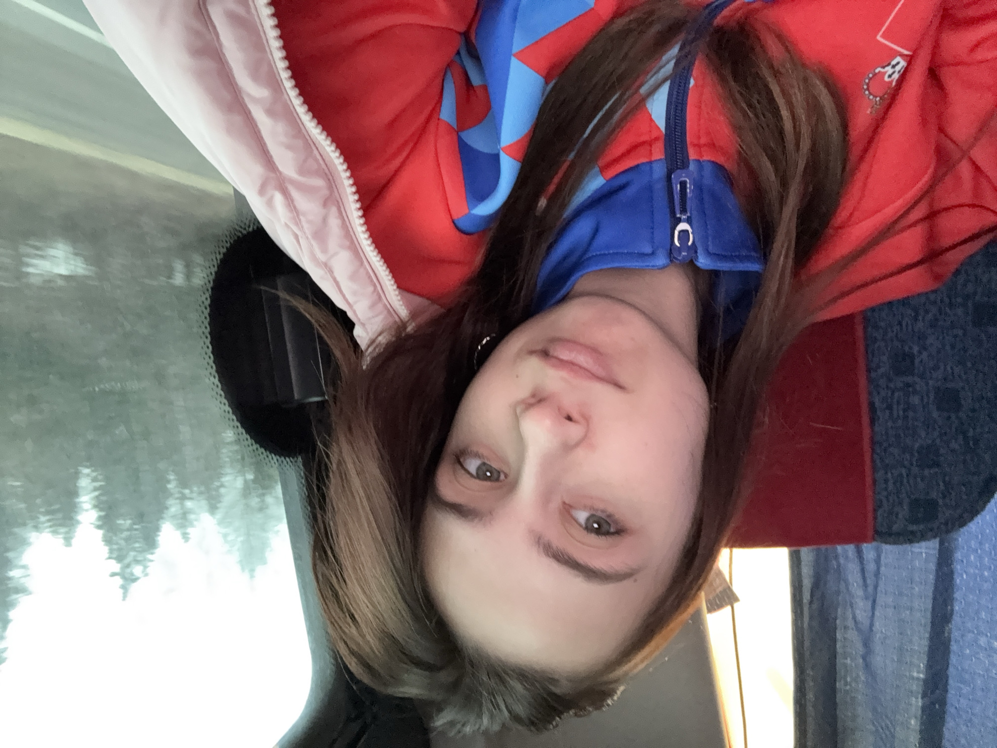 A young woman is looking at the camera, sitting inside a vehicle. She wears a red, blue, and pink jacket. Outside the vehicle is blurry, but appears to show trees in overcast conditions. There is no evidence to suggest this image was taken in Anninsk, Russia.